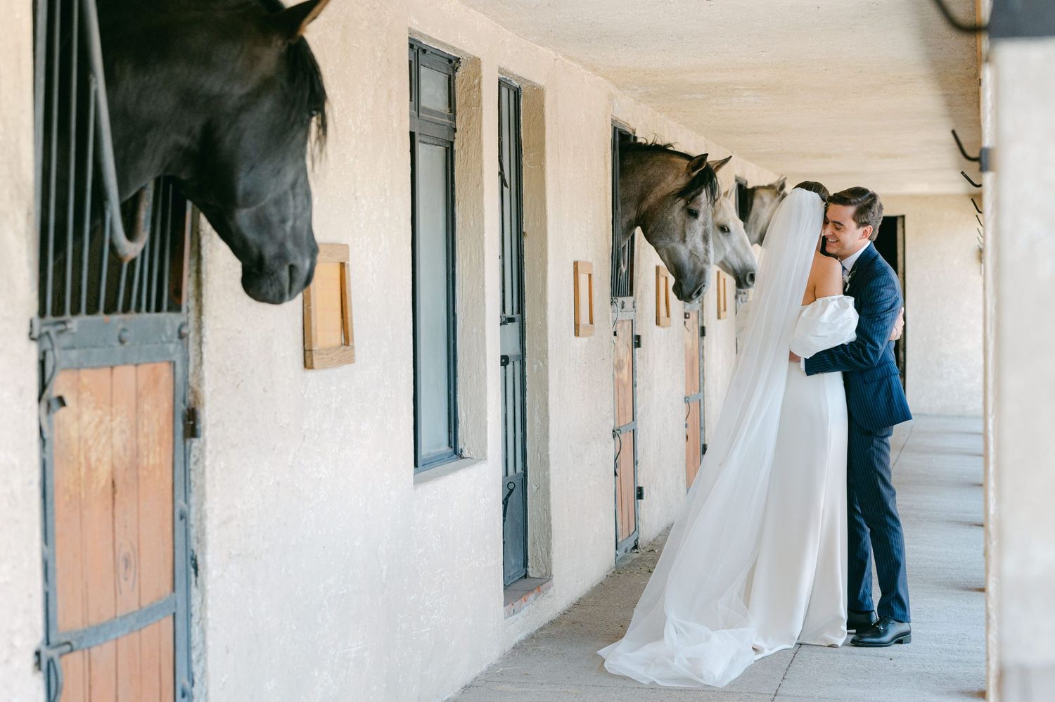 Bride and groom kiss in stable hallway as two horses lean out from their stalls nearby.