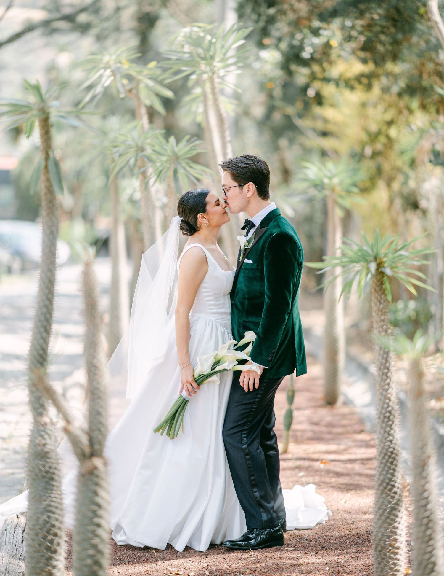 Bride and groom share a romantic kiss among tall palm trees, bride holding white calla lily bouquet wearing veil.