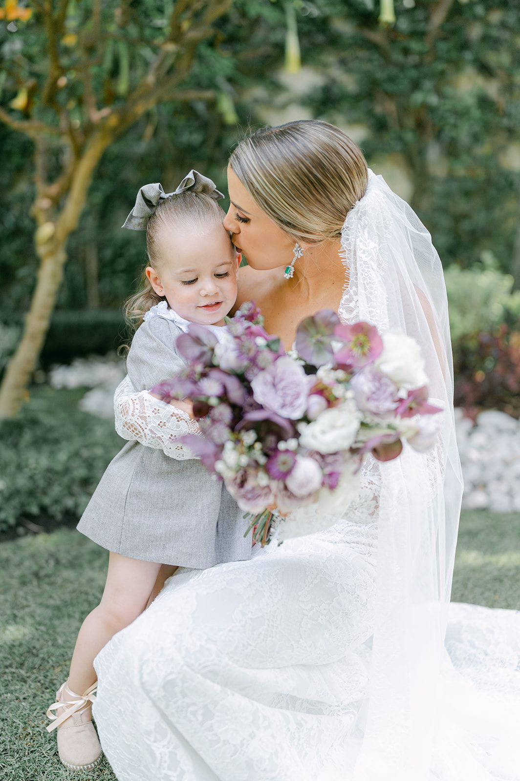 Bride in lace wedding dress kisses flower girl in gray dress while holding purple and white bouquet.