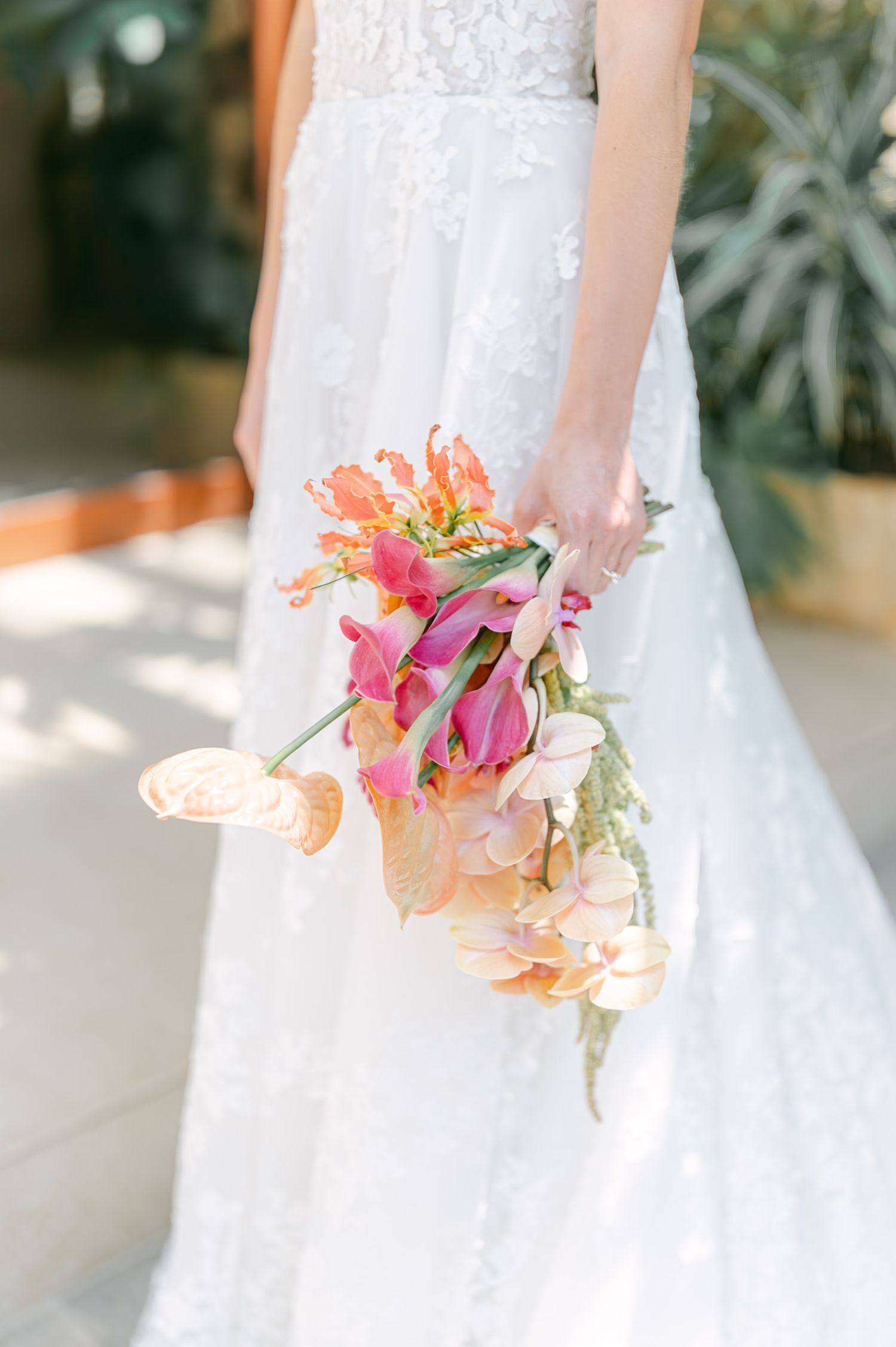 Close-up of bride holding vibrant tropical bouquet with orange anthuriums and pink flowers outdoors.