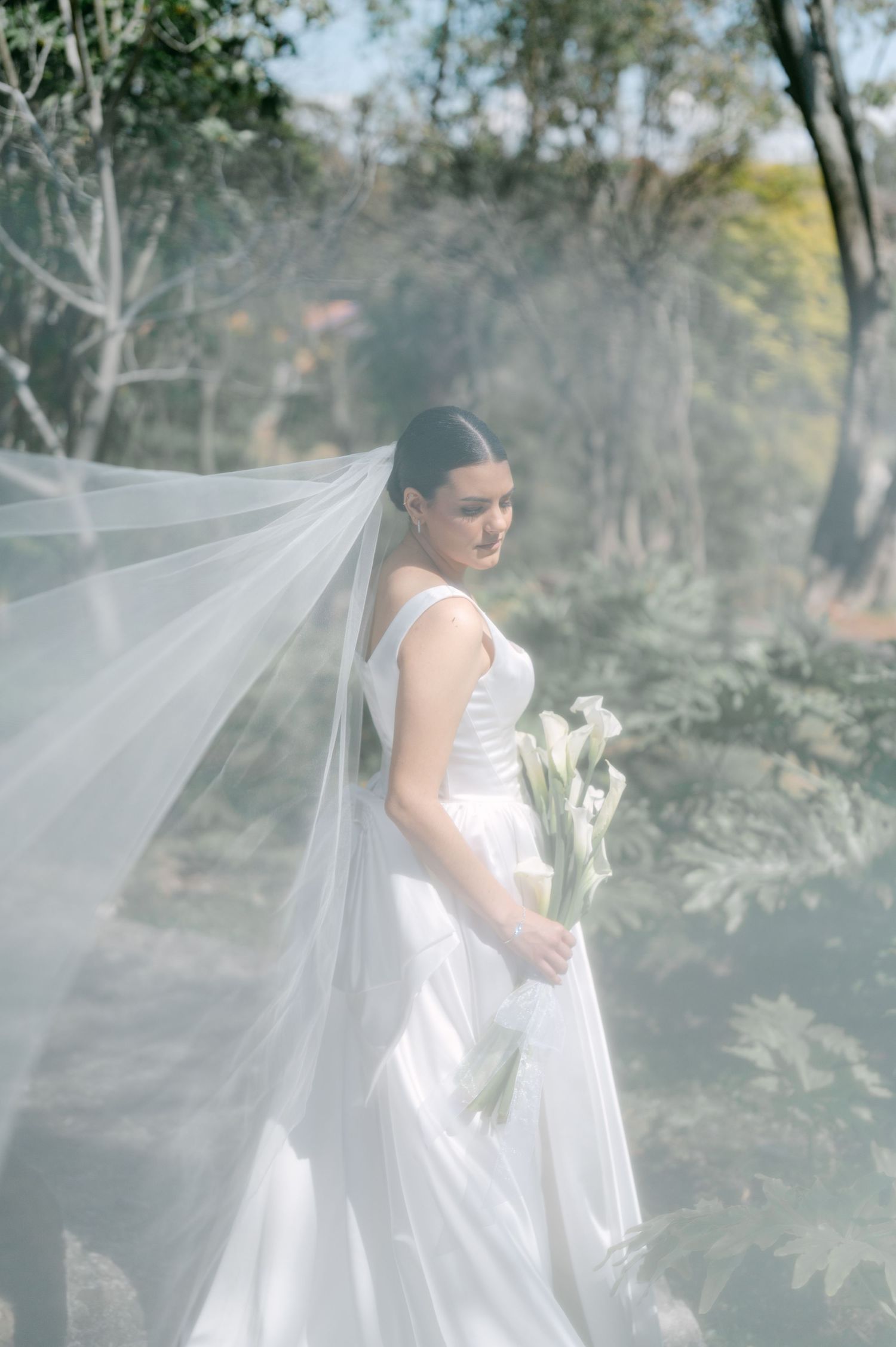 Bride in white gown holding calla lilies with flowing veil billowing in a sunlit woodland setting.