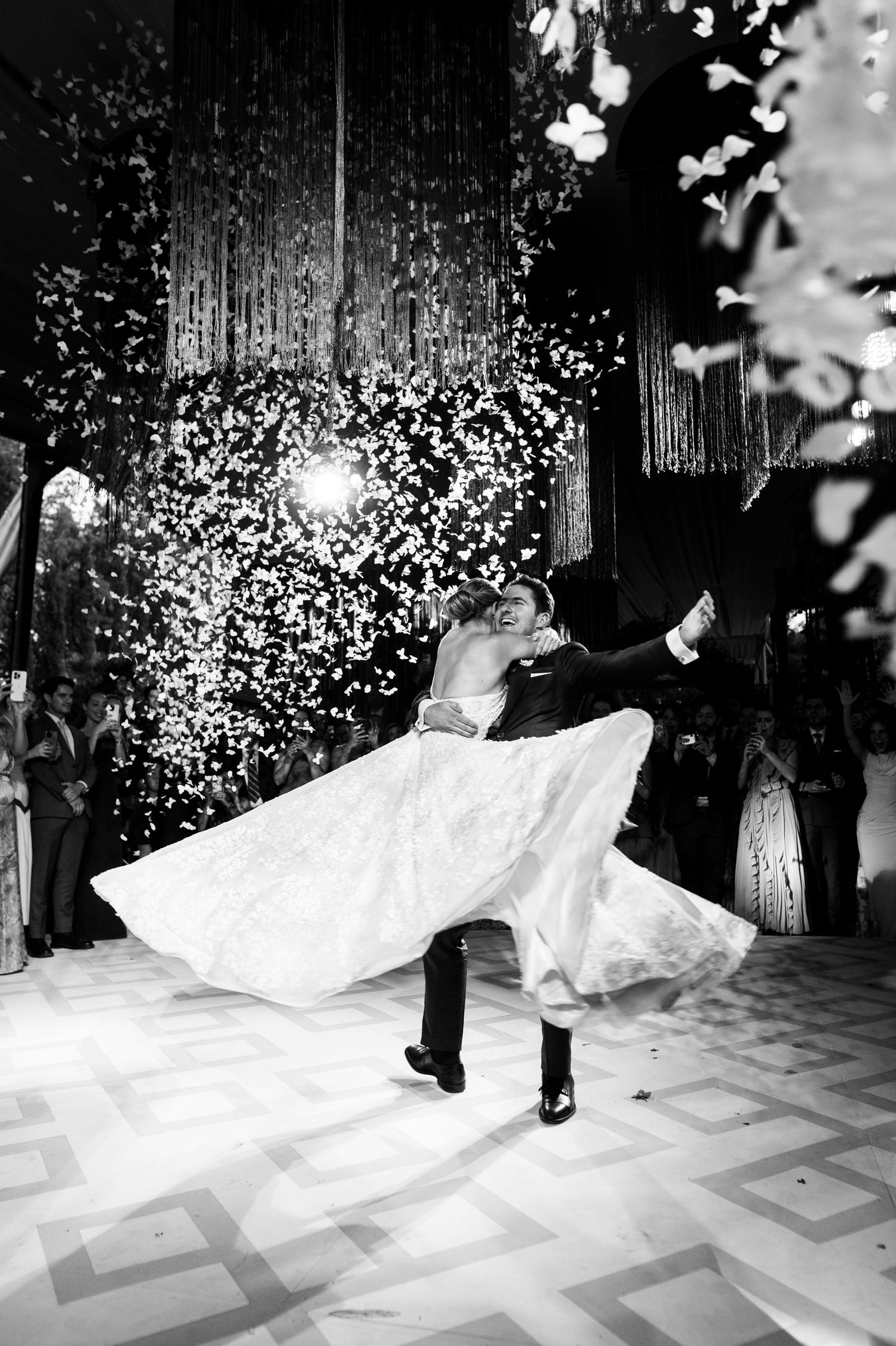 Black and white photo of a wedding couple dancing in an elegant ballroom with sparkling lights and decor.
