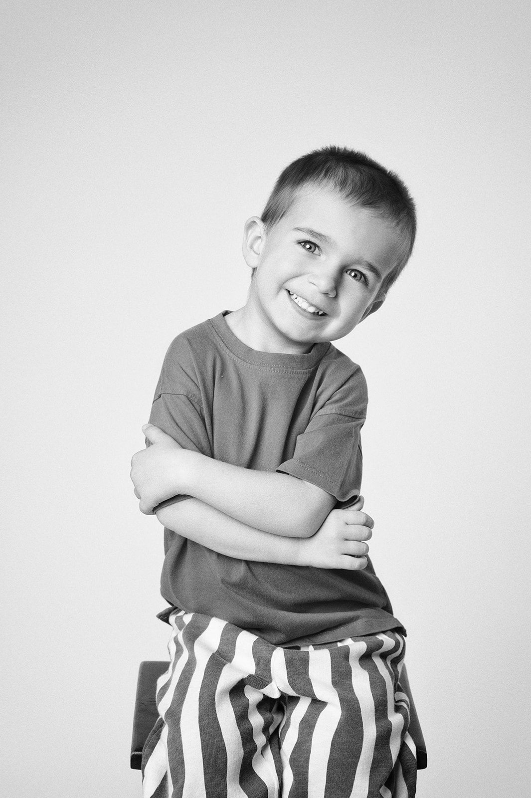 Black and white portrait of a smiling young boy with arms crossed, seated, wearing a striped pants and gray shirt.