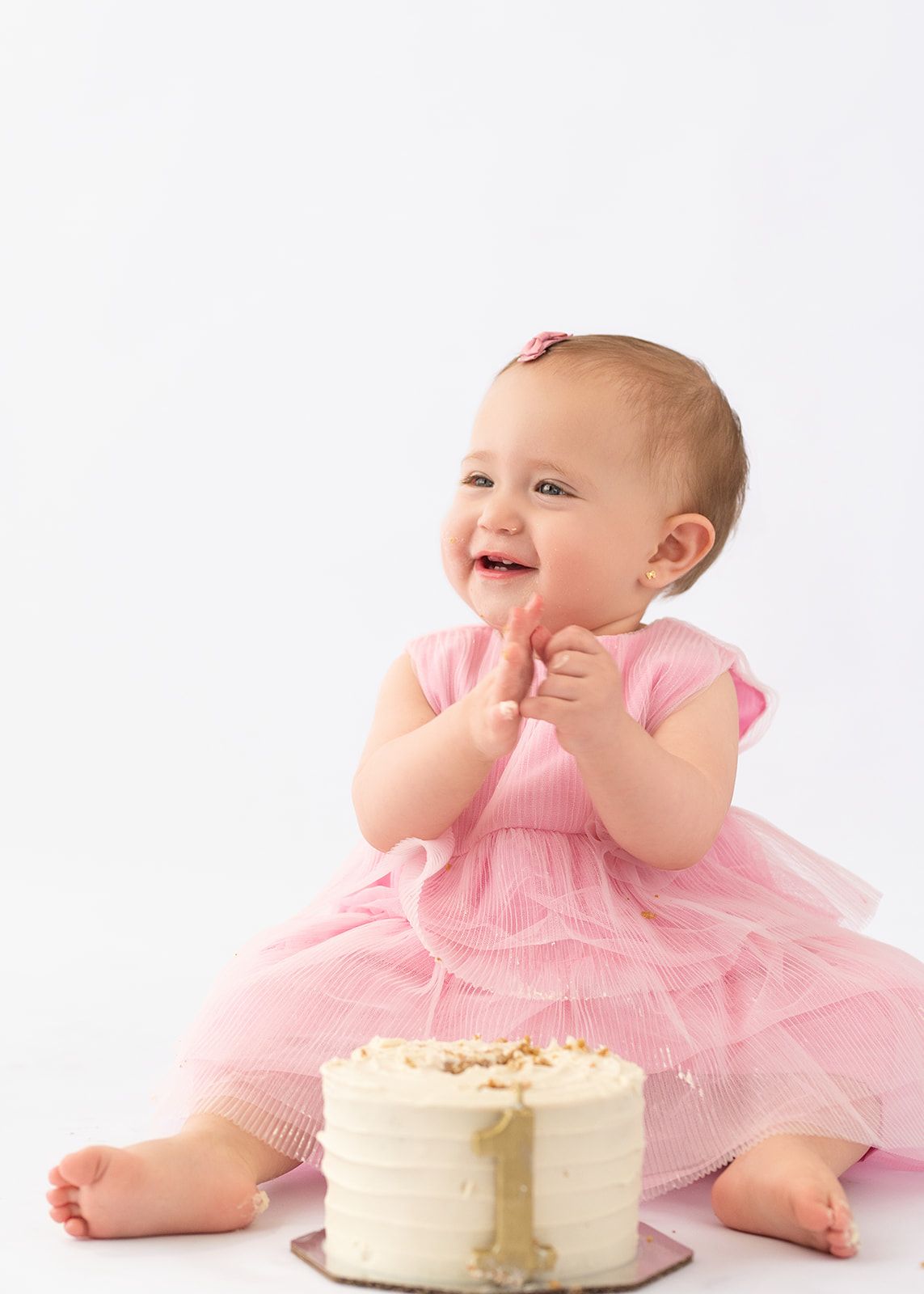 Smiling baby girl in pink tulle dress clapping hands beside a small white first birthday cake.