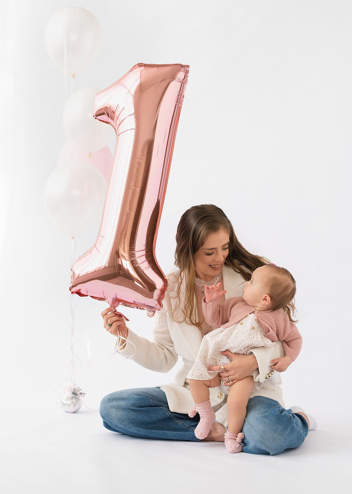 Mother holds baby girl while displaying a rose gold number one balloon at first birthday shoot.