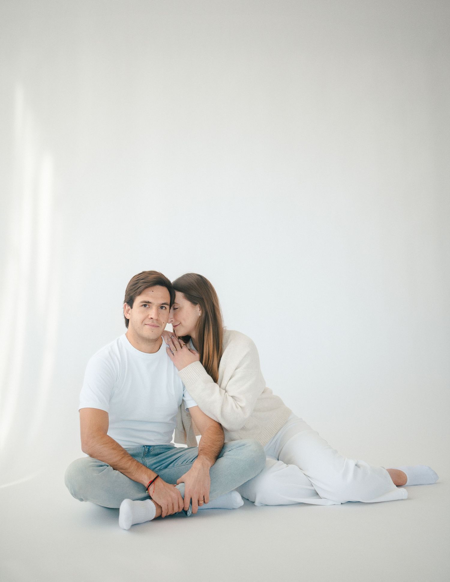 Couple sitting on a white studio floor with woman whispering to man in casual white and neutral outfits.