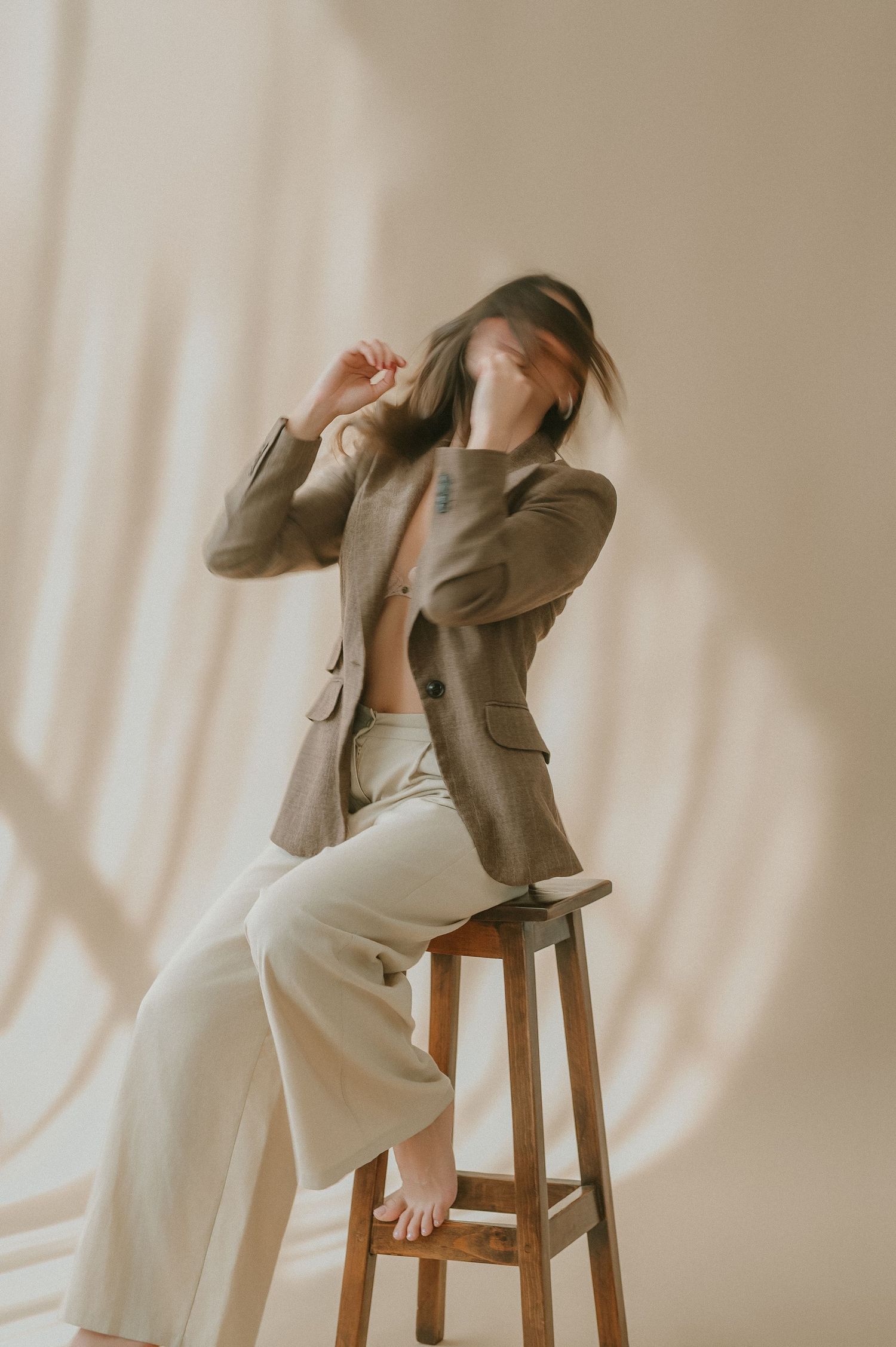 Woman in brown blazer with open shirt sits on wooden stool, hair blurred in motion, soft shadow background.