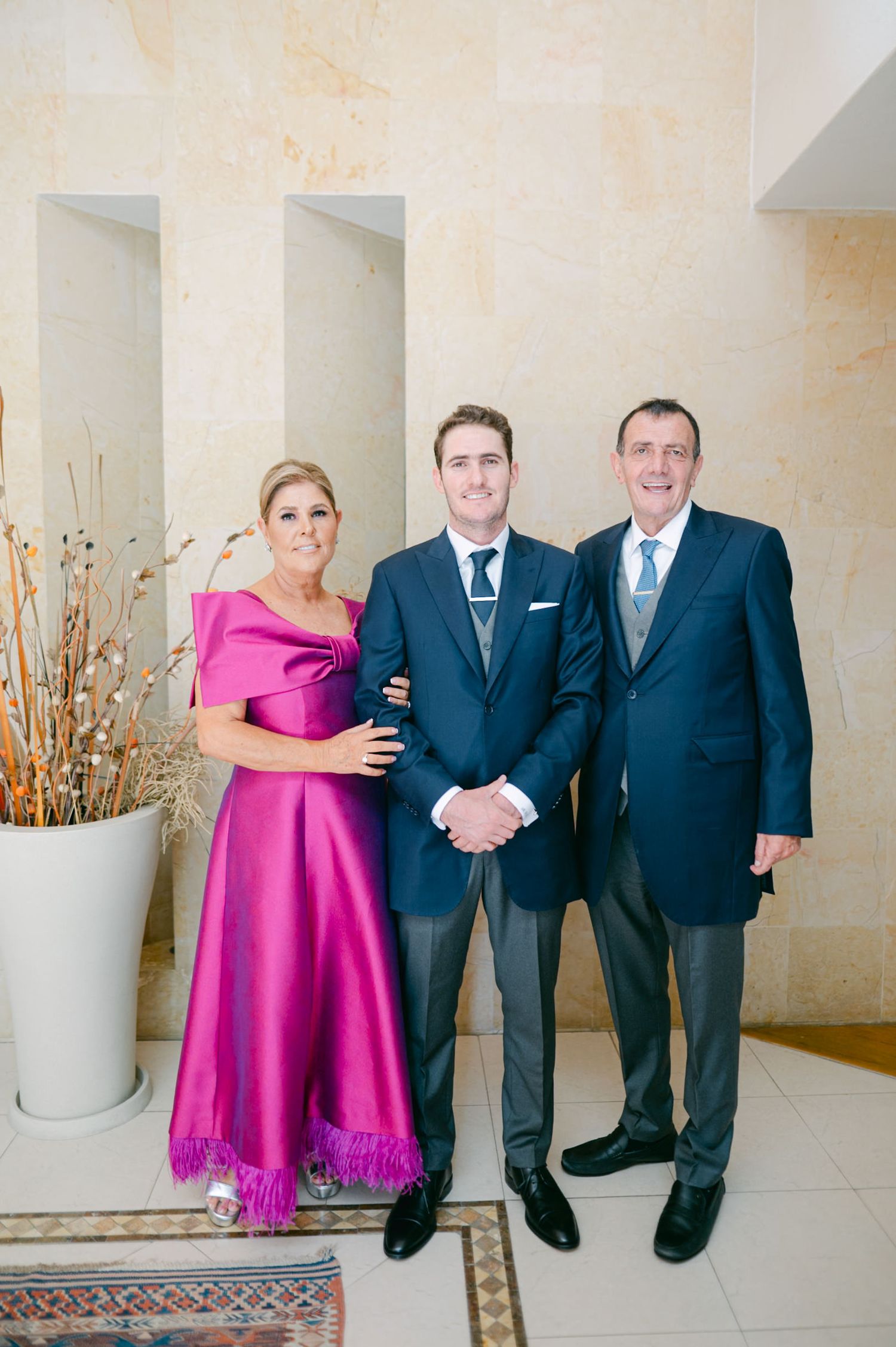 Groom posing with parents, mother in fuchsia gown and father in navy suit, before wedding.