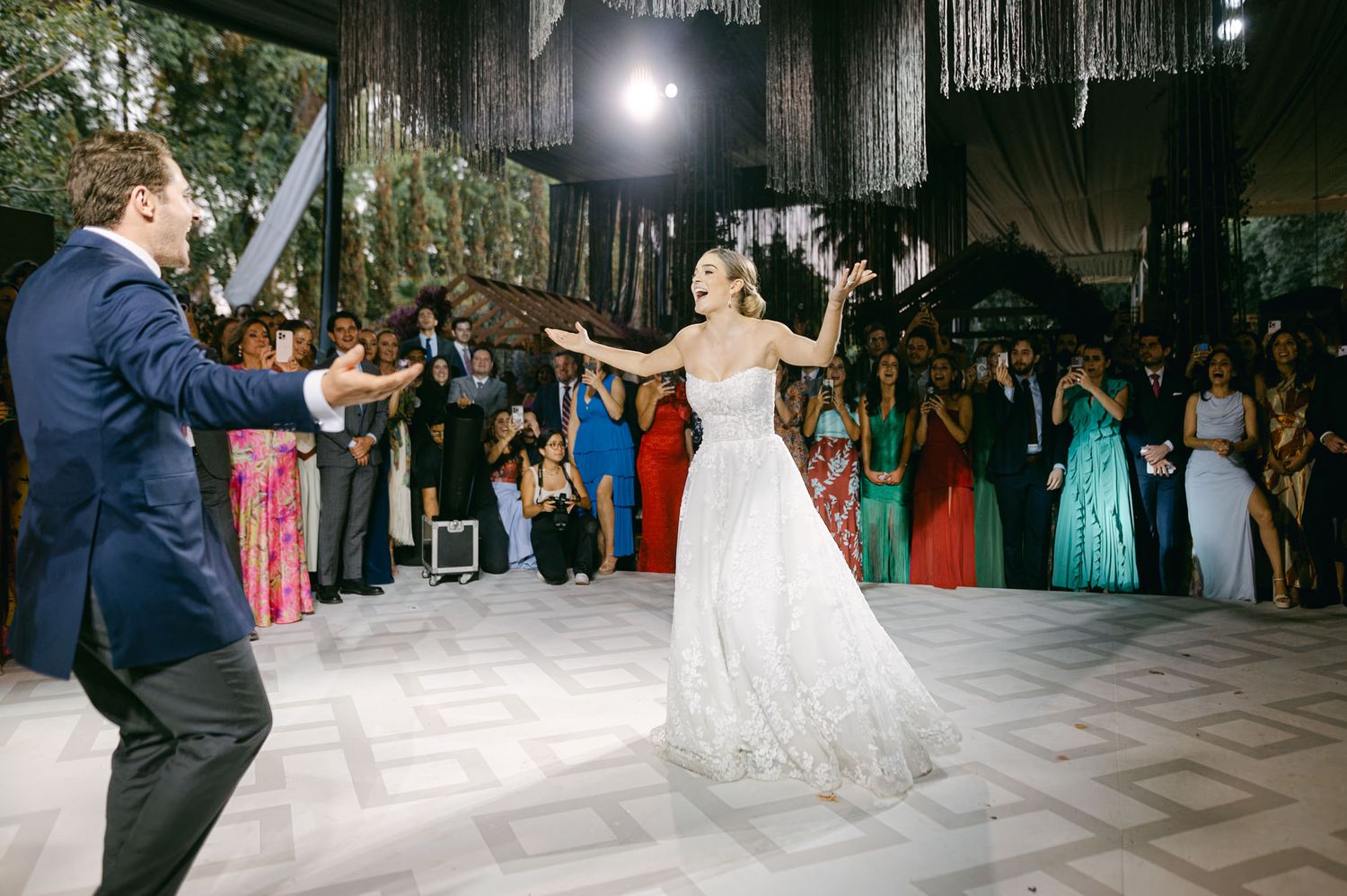 Bride and groom dance enthusiastically during wedding reception under hanging decorations.