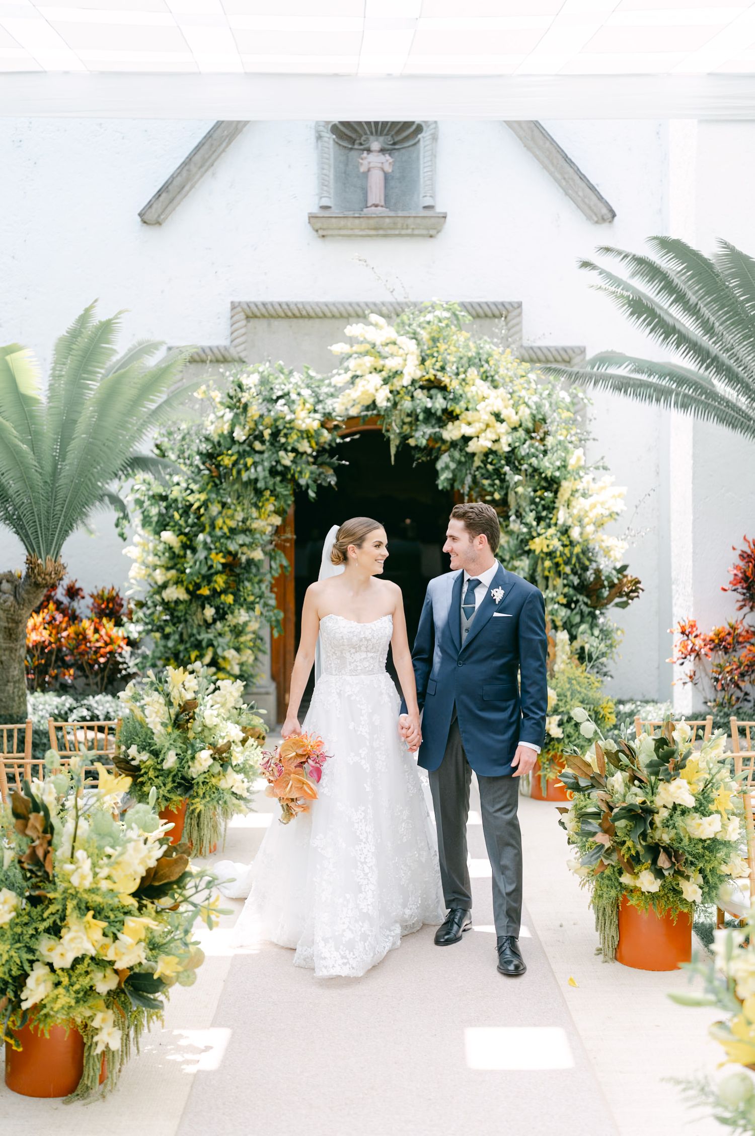 Bride and groom exit church ceremony through an elaborate floral arch with colorful tropical blooms.