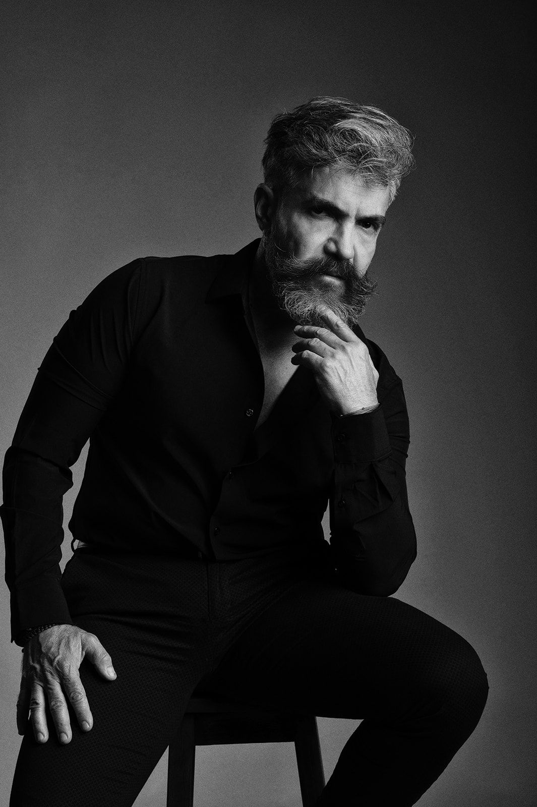 Black and white portrait of a bearded man in black shirt sitting on stool with thoughtful pose.