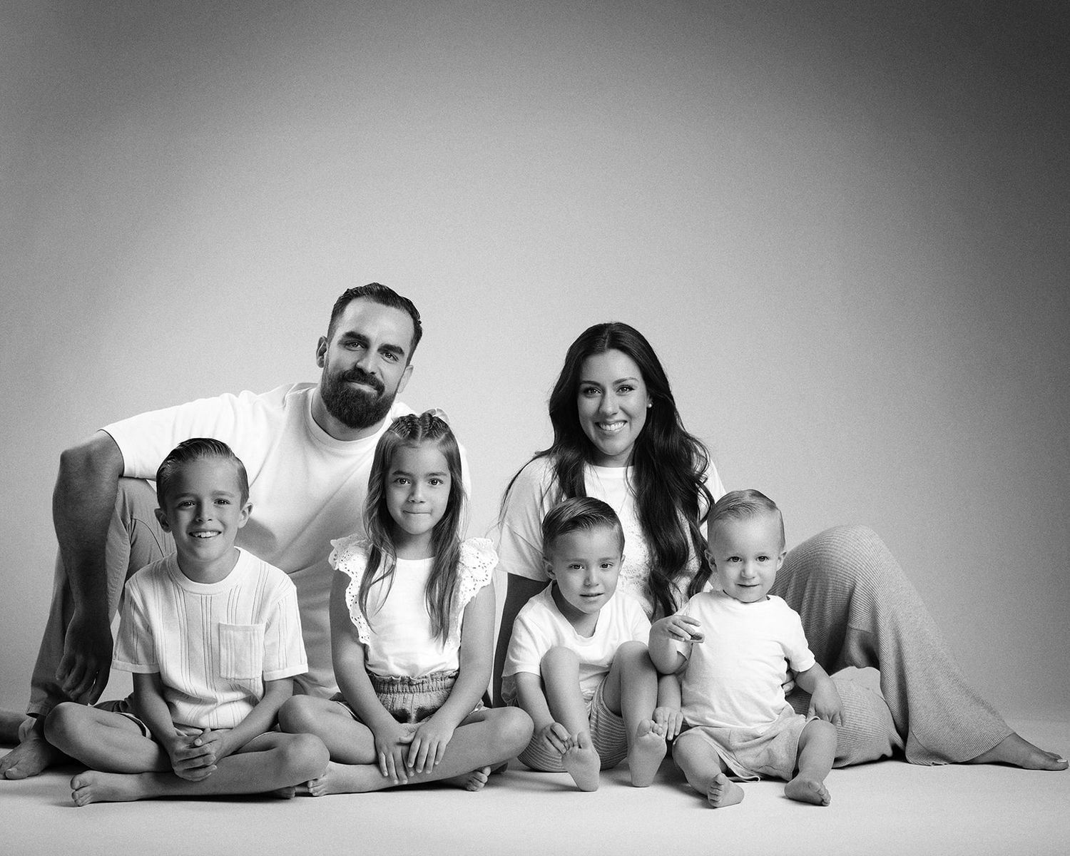 Black and white studio portrait of a family of six sitting together on the floor, smiling at the camera.