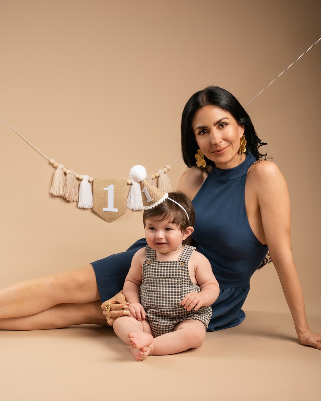 Mother and baby girl pose together at a first birthday photoshoot with a boho tassel number one banner.