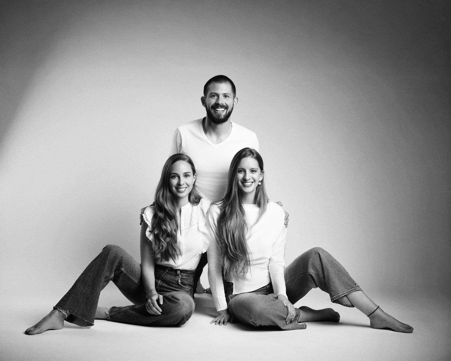 Black and white studio portrait of a man and two women sitting together on the floor smiling.