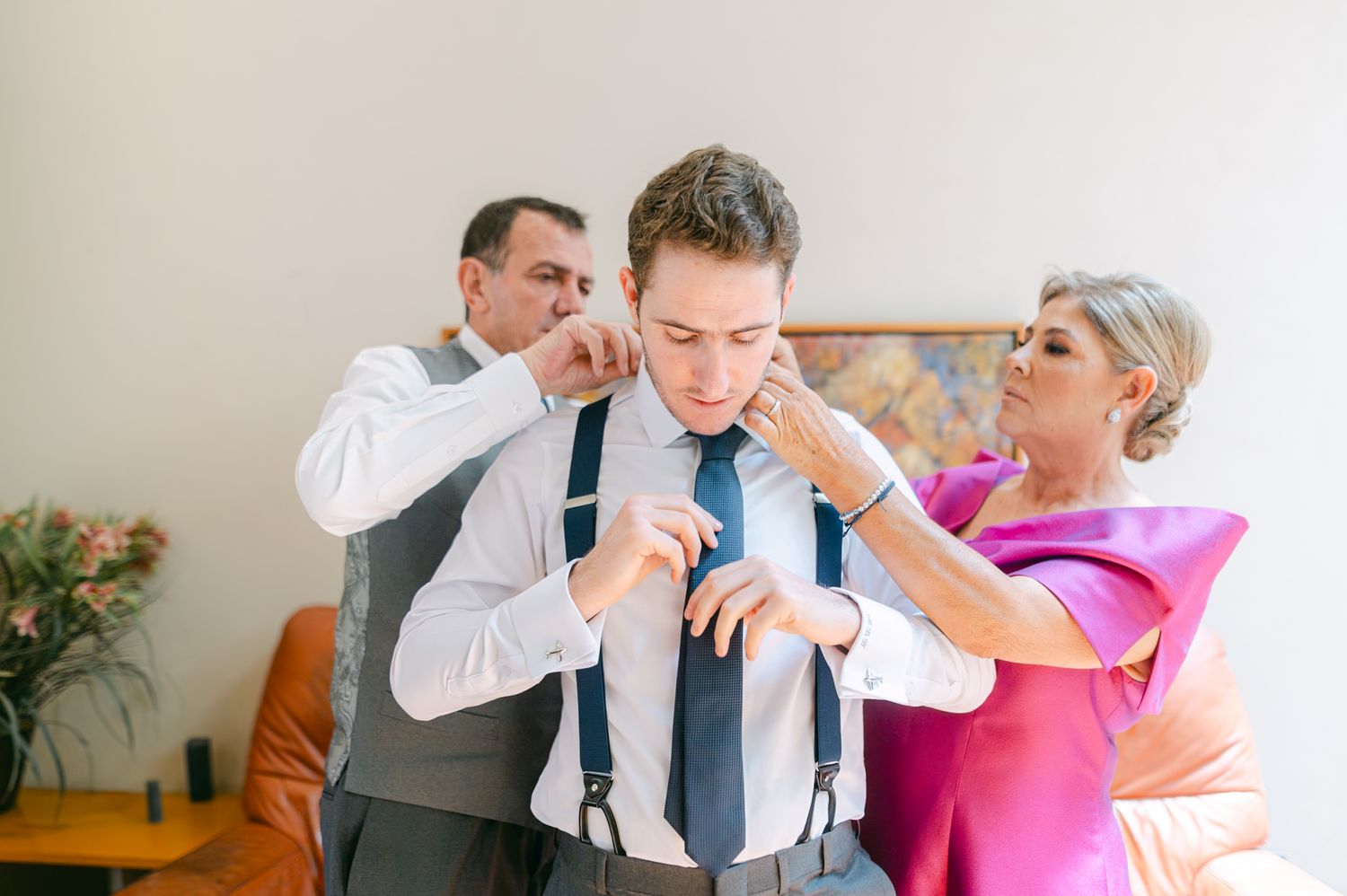 A groom in suspenders gets ready as his parents help him with his tie and collar.