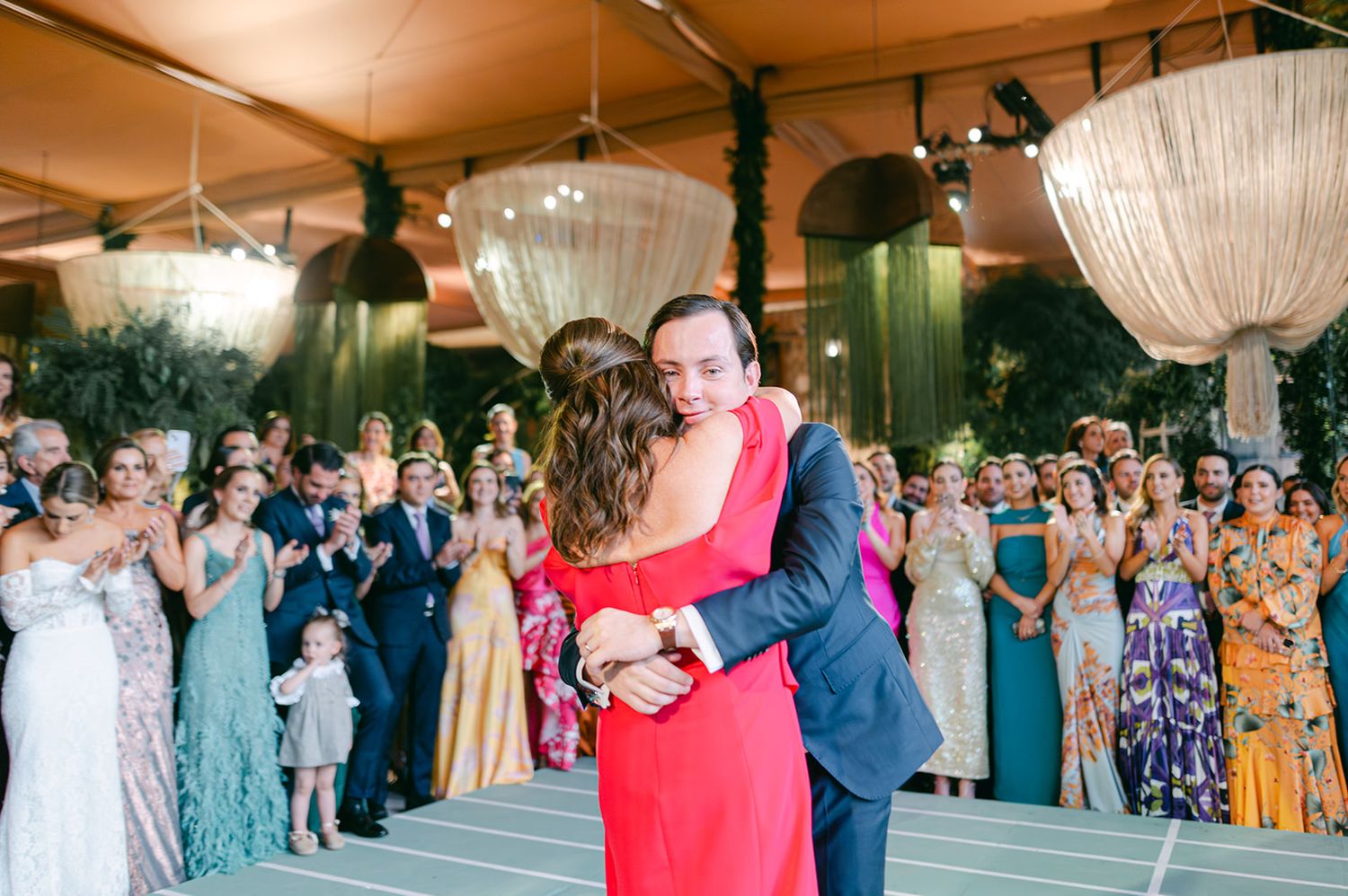 Groom in navy suit shares warm embrace with woman in red dress on wedding reception dance floor.