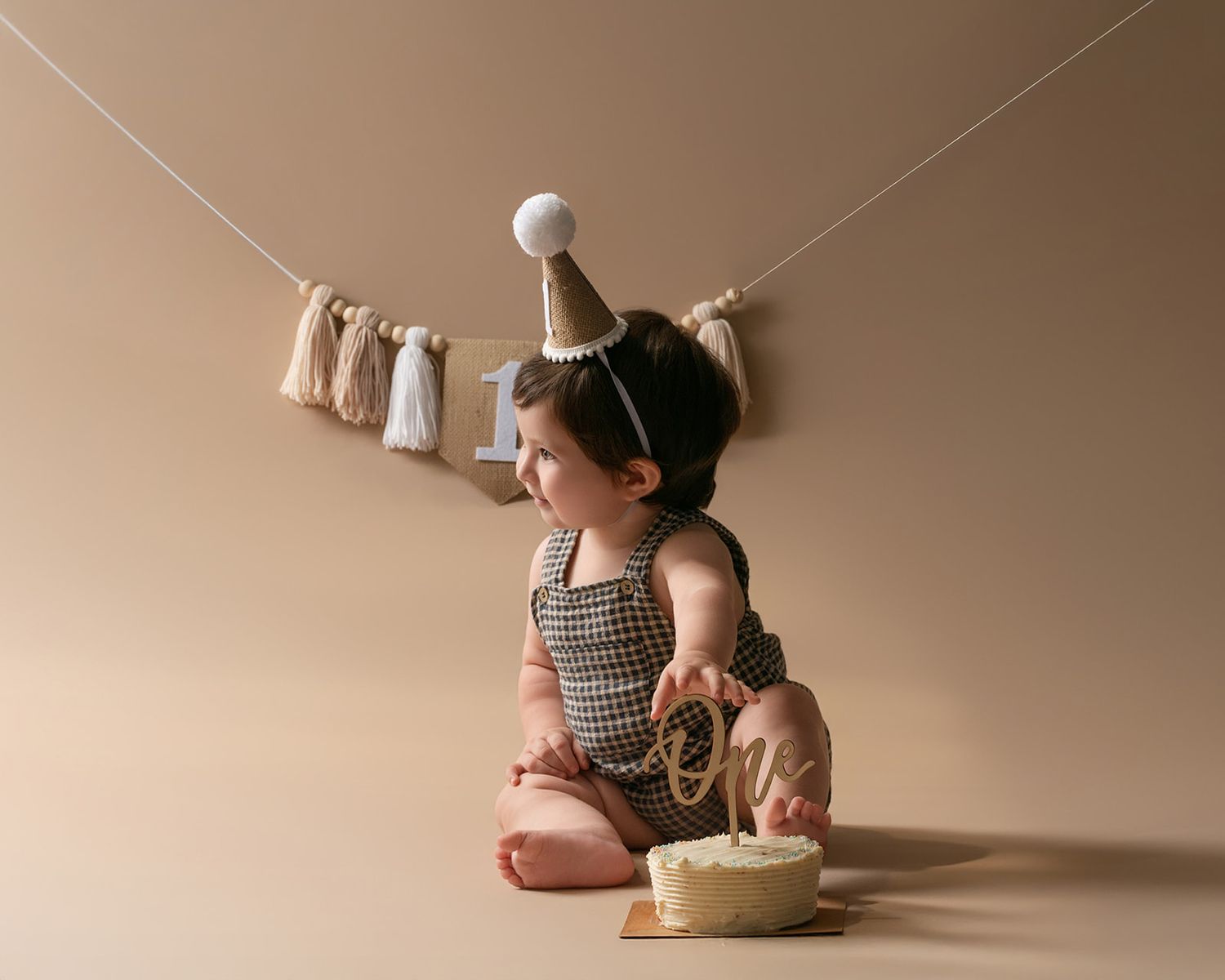 A baby in a checkered romper and party hat smashes their first birthday cake in a studio setting.