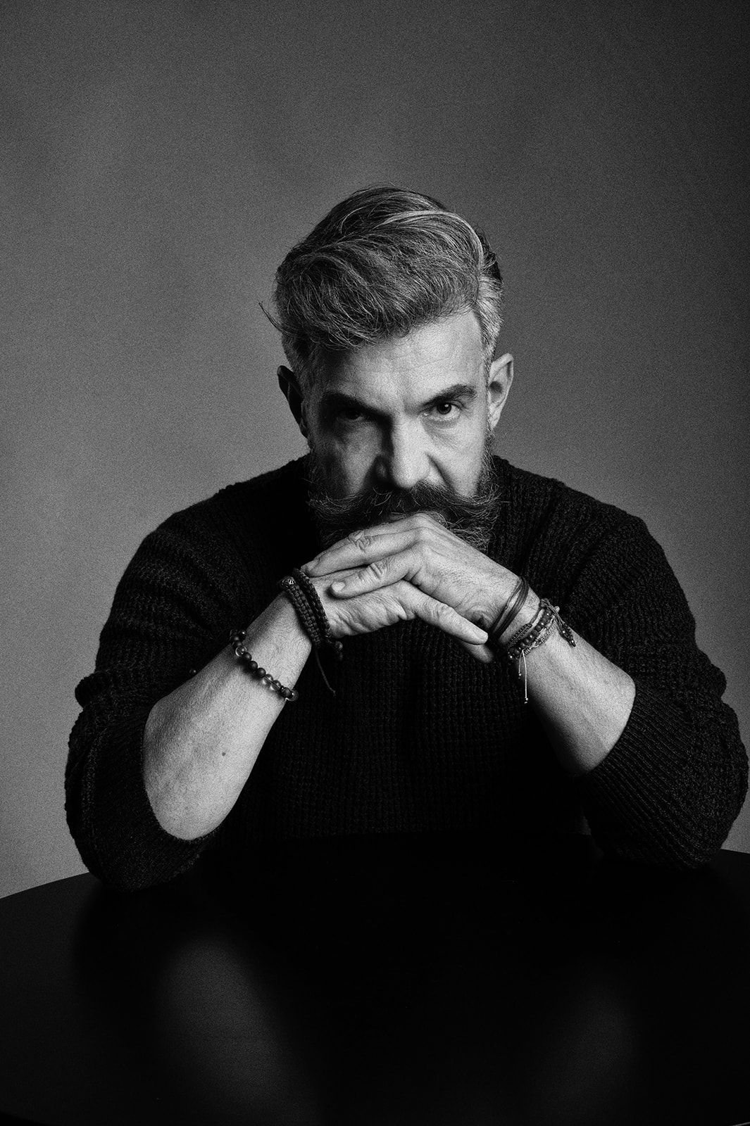 Black and white studio portrait of a bearded man in dark sweater with hands clasped at table.