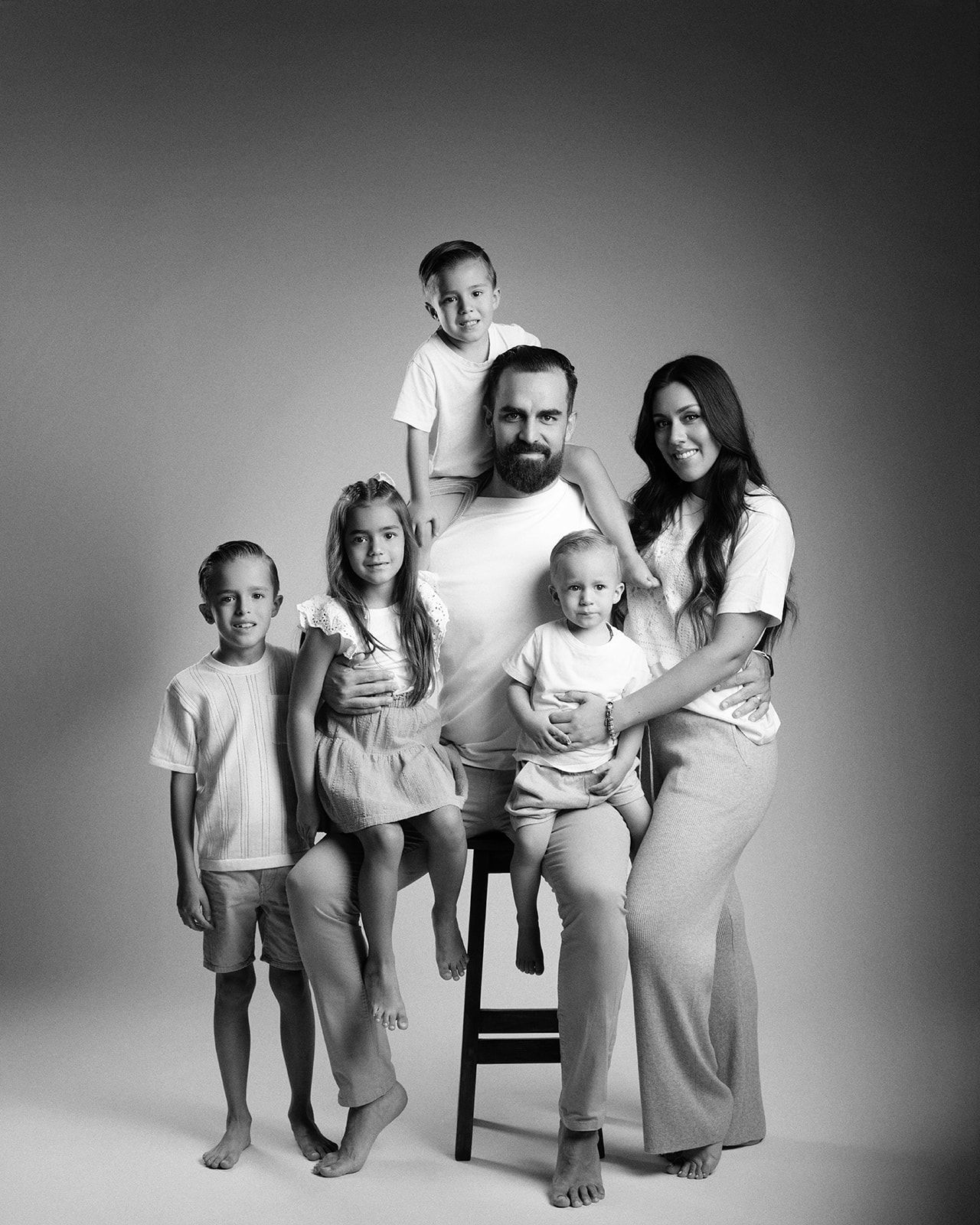 Black and white family portrait with bearded father, mother, and four children posing with a stool in a studio.