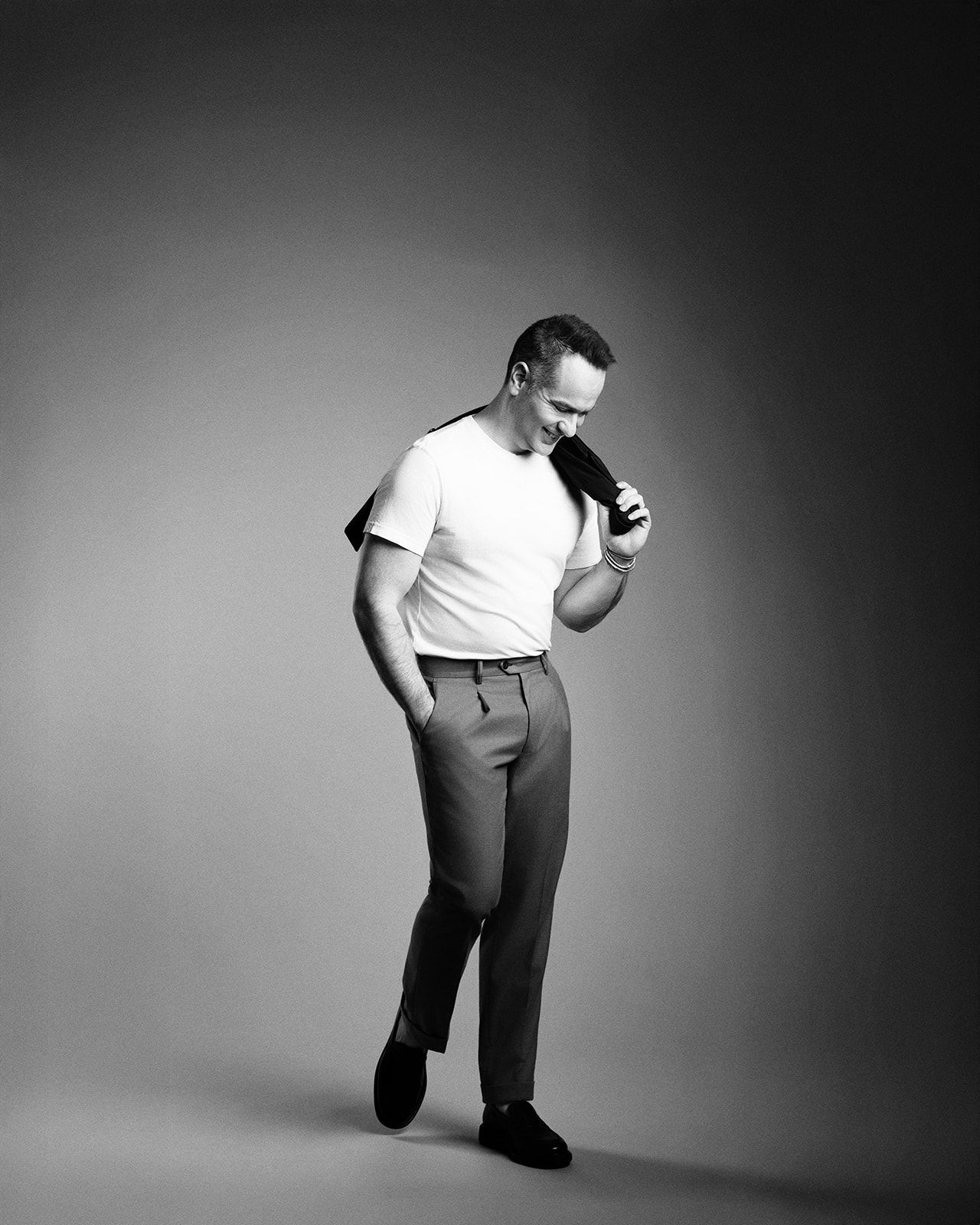 Black and white photo of a man in white t-shirt and trousers holding a jacket, looking down in a studio setting.
