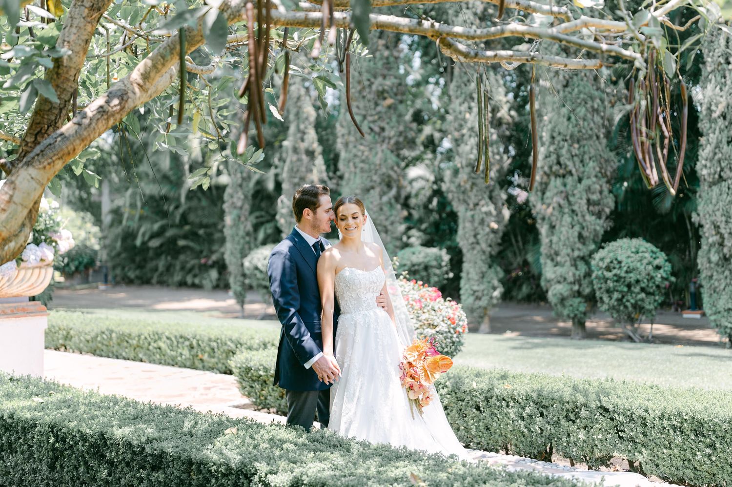 Bride and groom share a romantic moment among lush garden hedges, bride holding a vibrant bouquet.