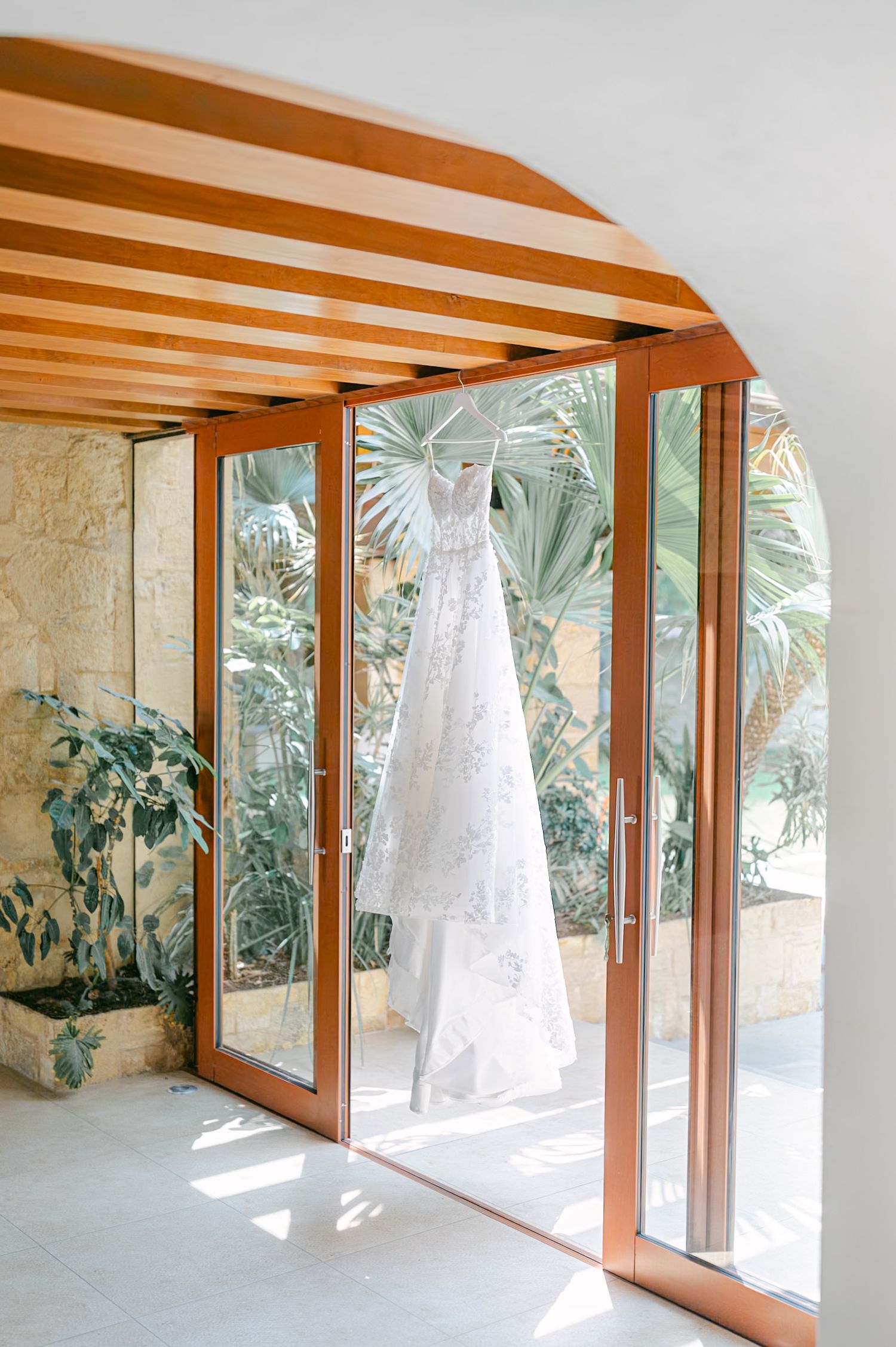 A lace wedding dress hanging on a hanger in a sunlit arched wooden doorway with tropical plants.