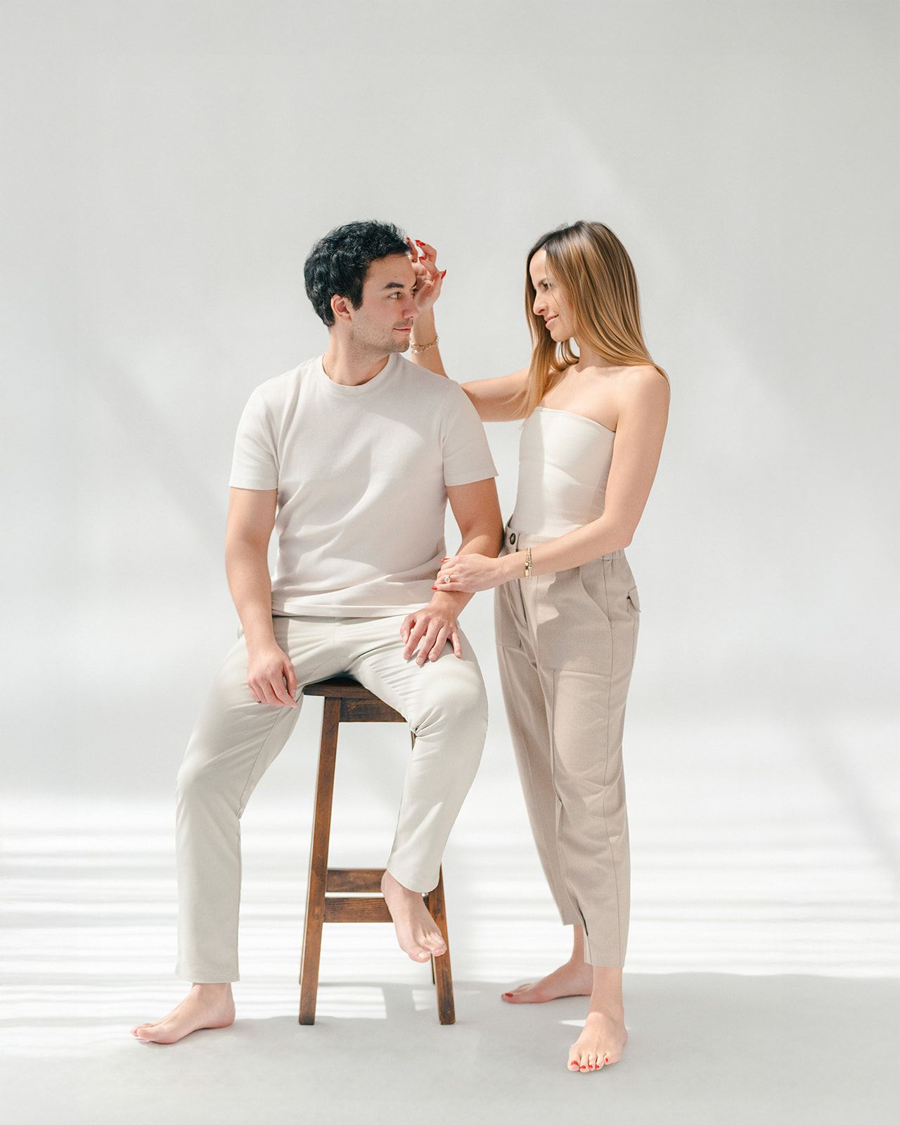 Man sitting on wooden stool with woman standing beside him in coordinated beige casual outfits on white background.