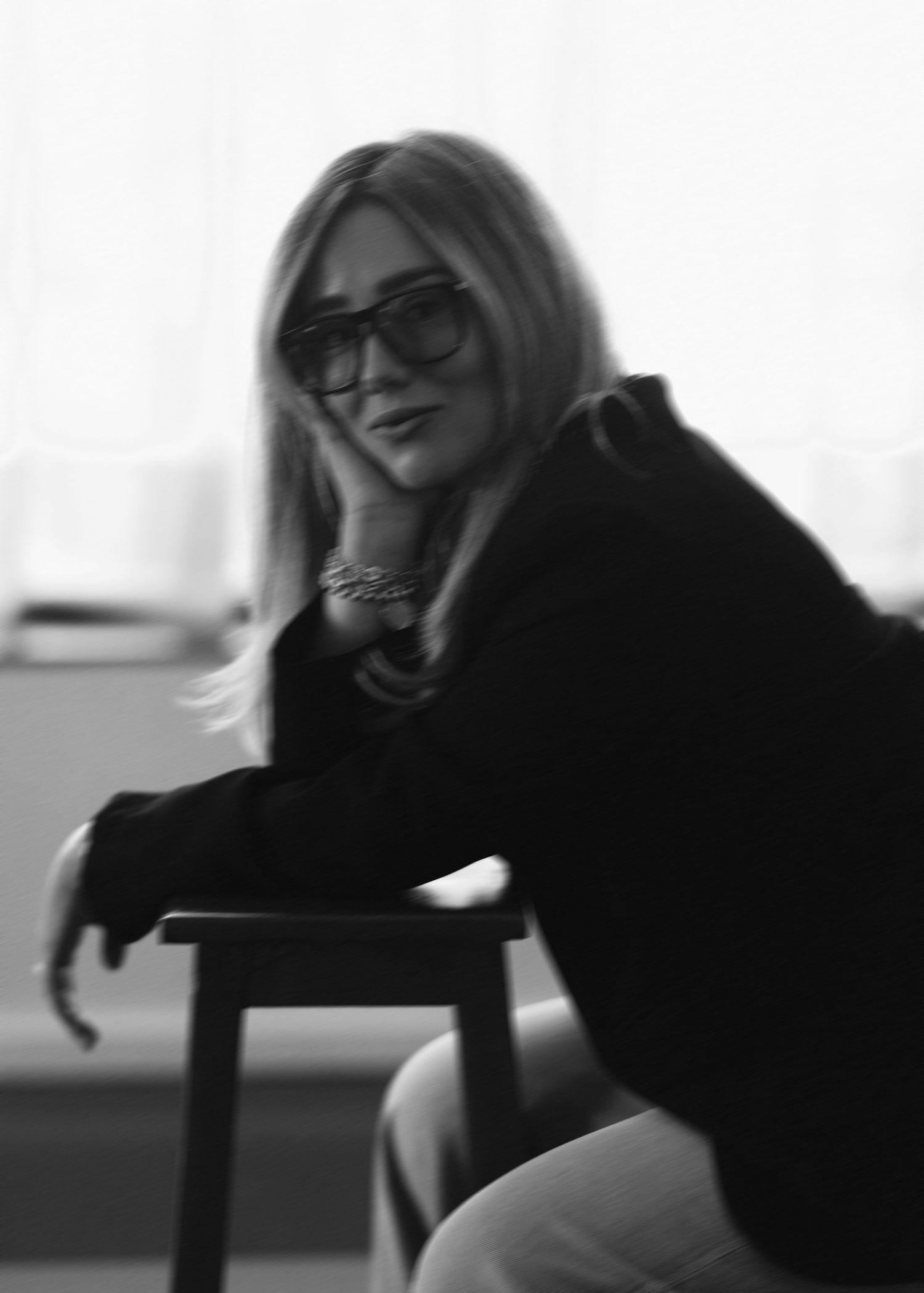 Black and white photo of a blonde woman in sunglasses seated on a stool in soft light.