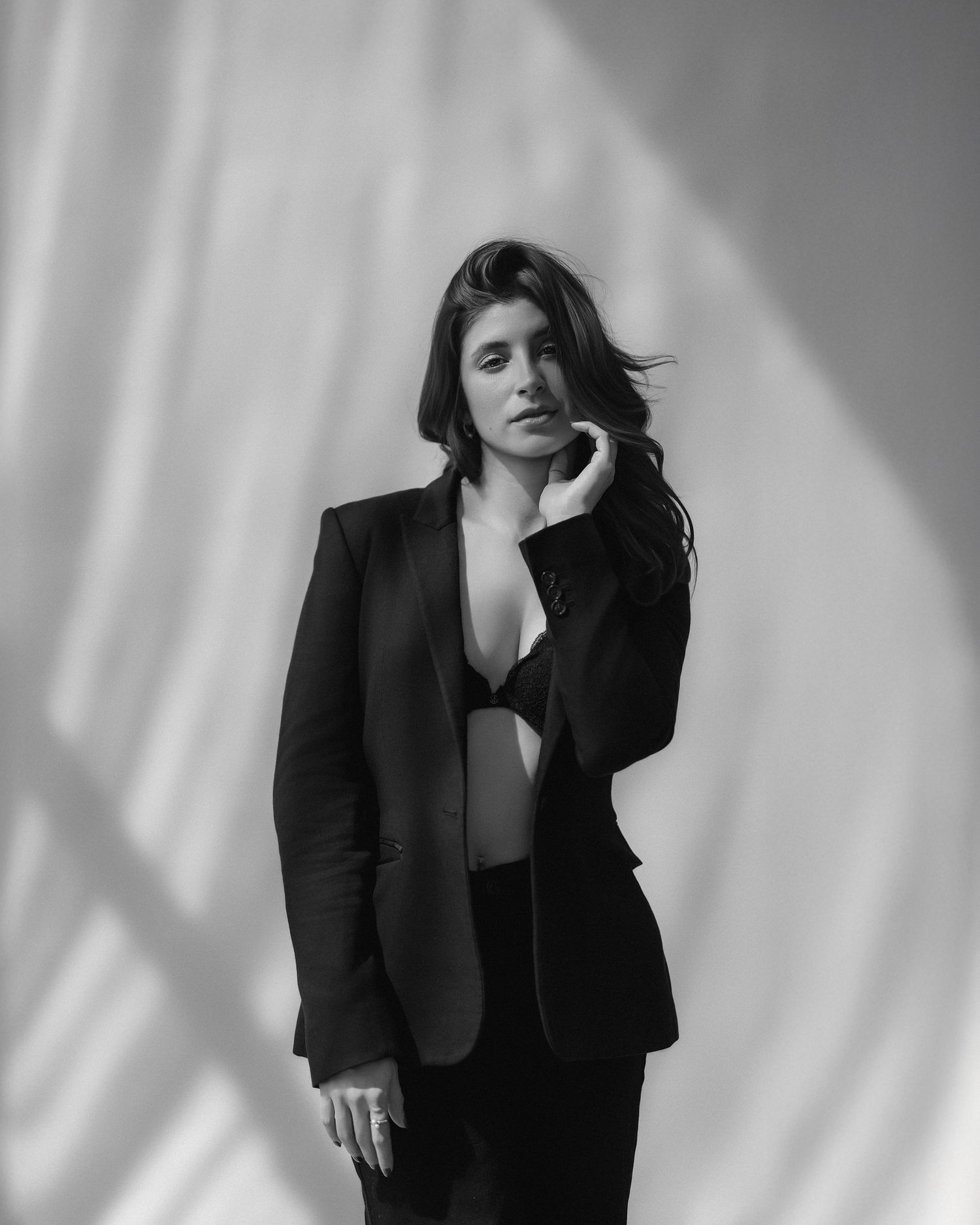 Woman in black blazer and lingerie poses confidently, black and white studio portrait with soft shadows.
