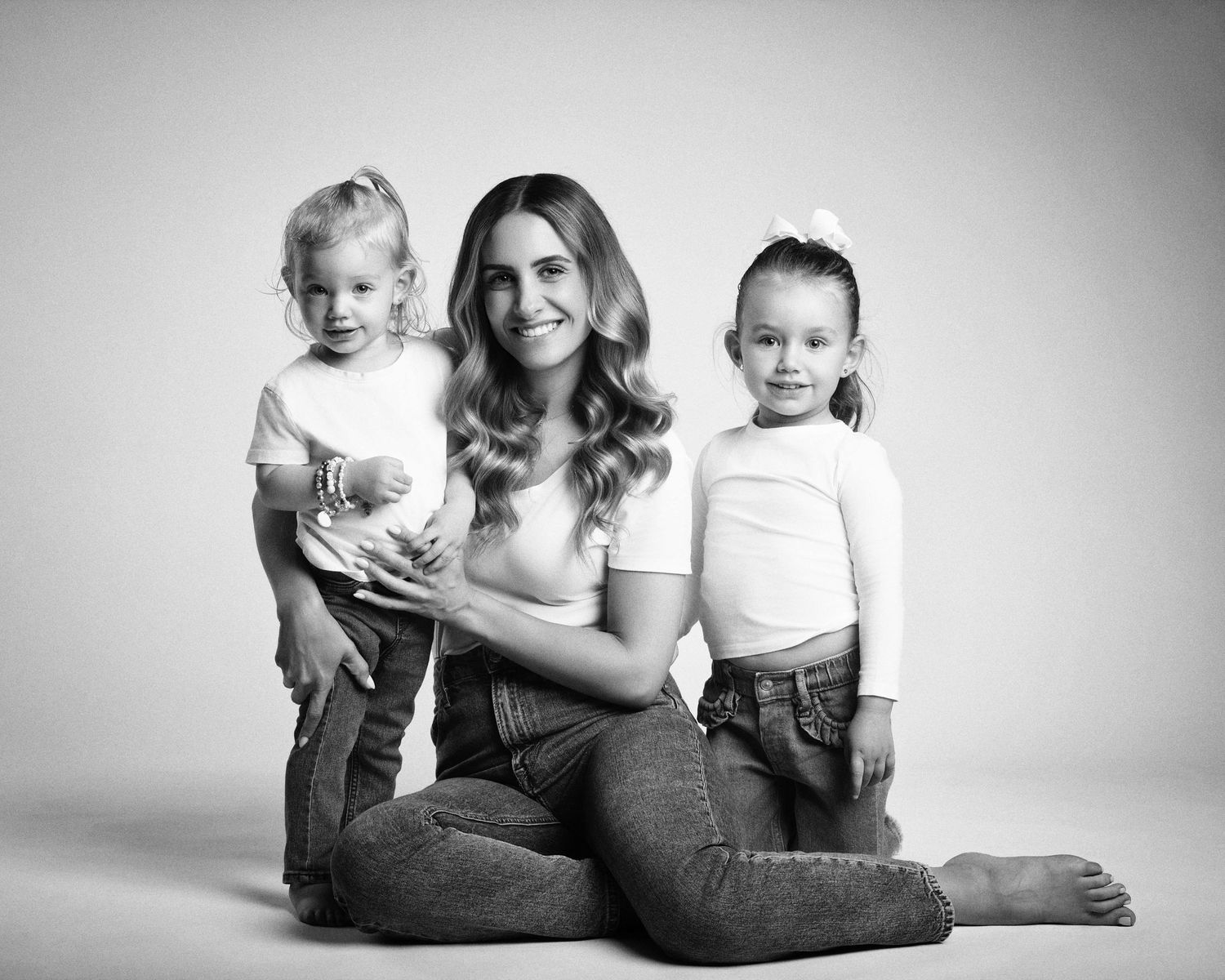 Black and white portrait of a mother sitting on the floor with her two young daughters smiling.