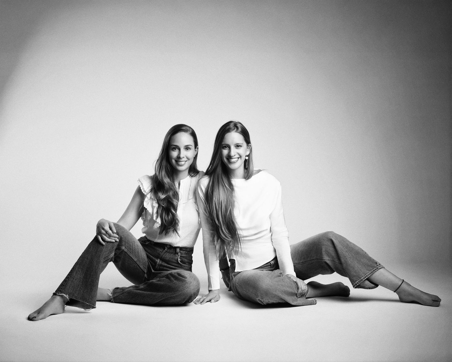 Two young women sit together on the floor in a black and white studio portrait wearing jeans.