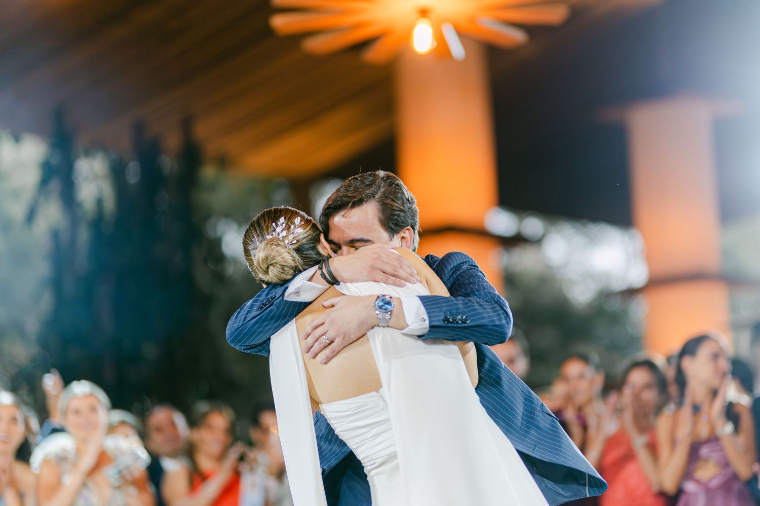 Bride and groom share a tender embrace on the dance floor surrounded by cheering wedding guests.
