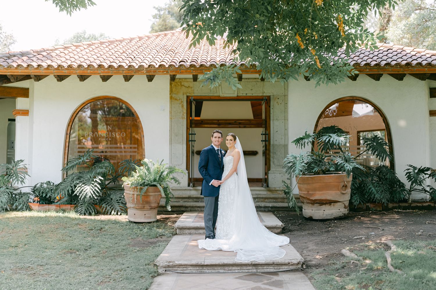 Bride and groom posing together in front of white Spanish-style wedding venue with arched windows.