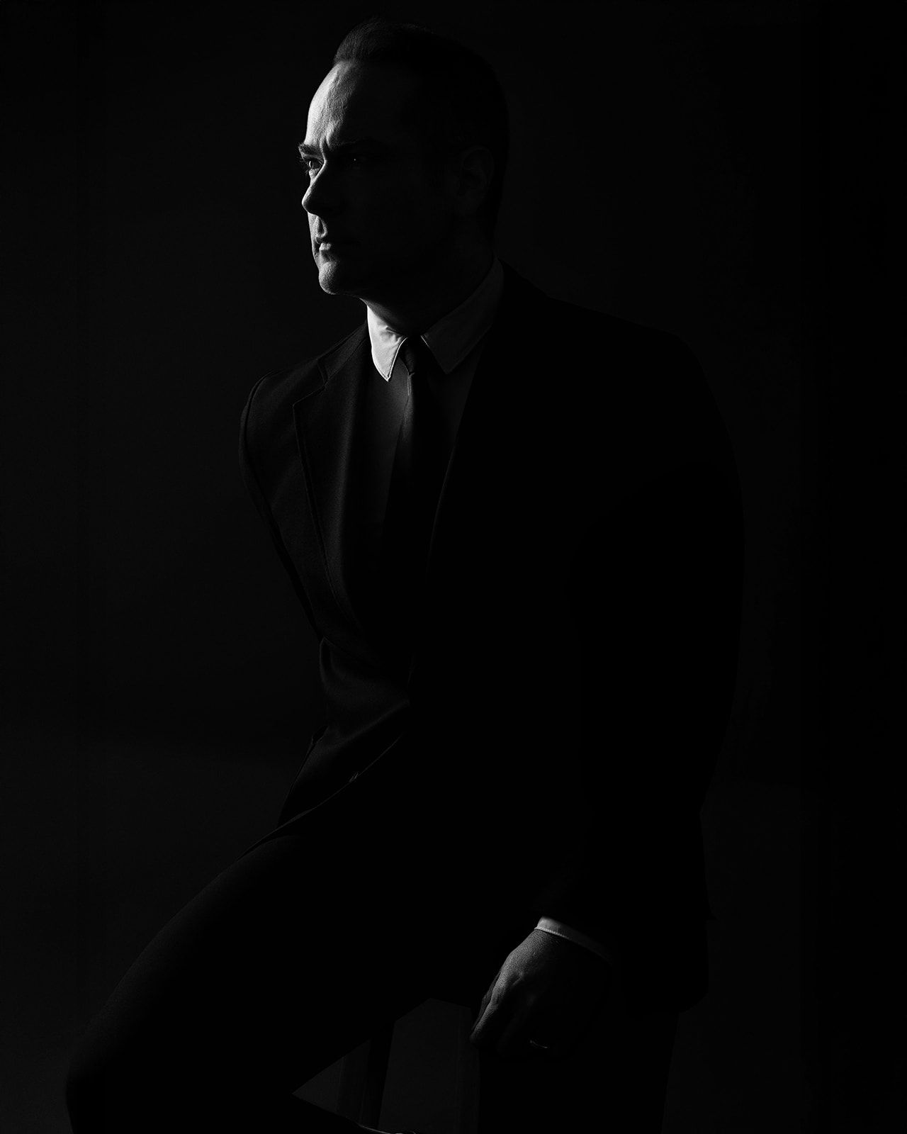 Dramatic black and white silhouette portrait of a man in a dark suit against a moody dark background.