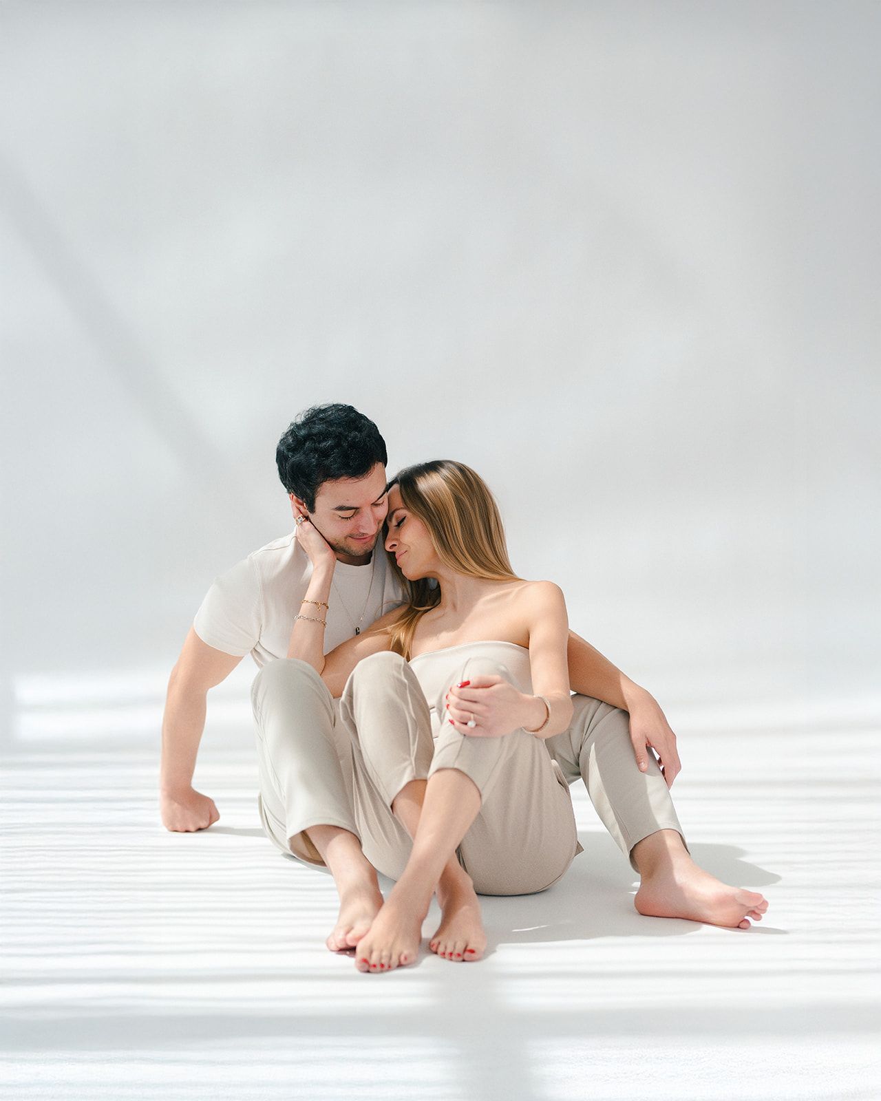 A couple in neutral tones sits intimately on a white floor, sharing a tender moment in soft natural light.