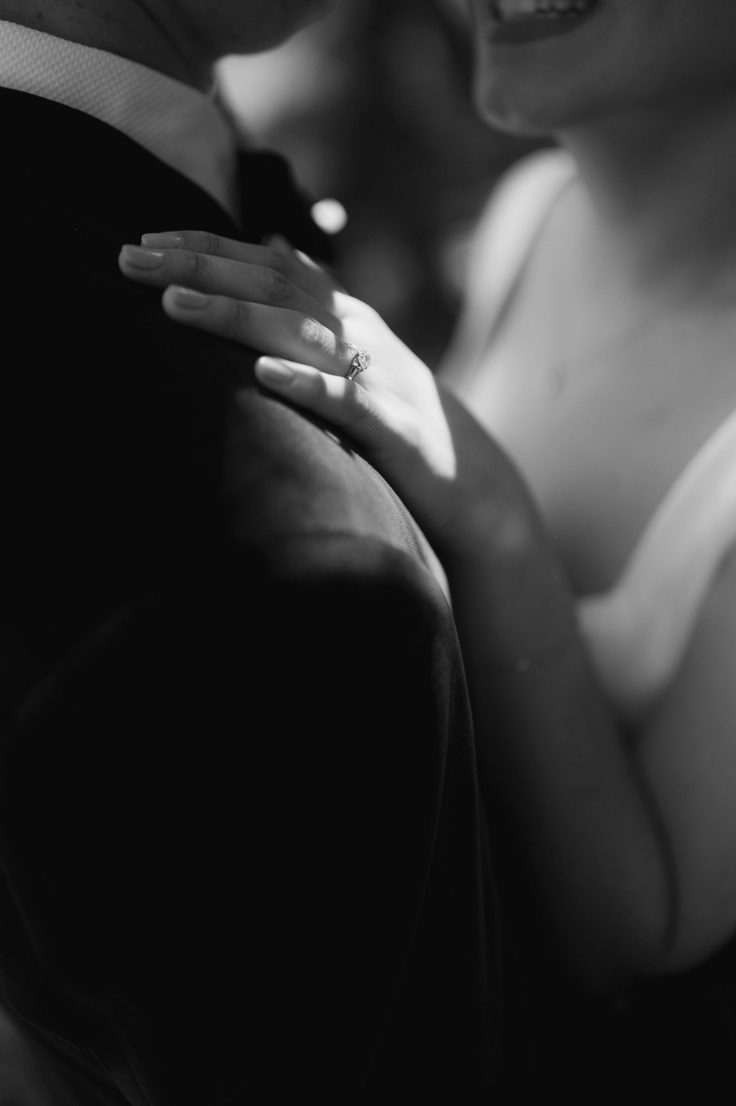 Black and white close-up of a bride's hand with engagement ring resting on groom's shoulder.