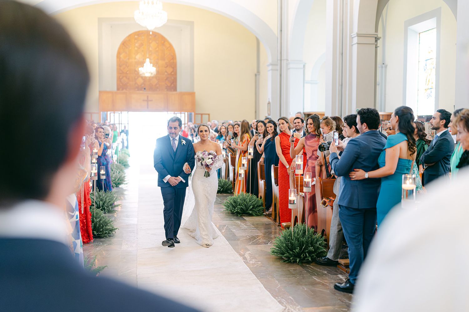 Bride in white lace gown walks down church aisle with escort past rows of wedding guests.