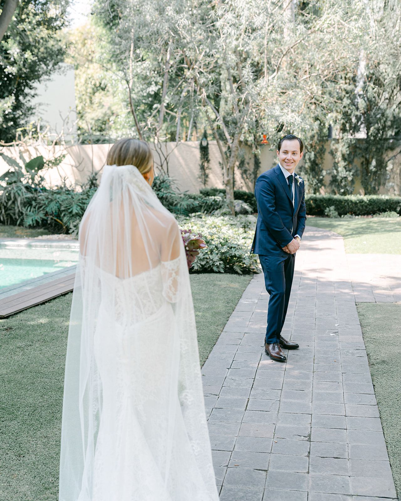 Groom in navy suit smiling at bride in cathedral veil during outdoor first look by a pool.
