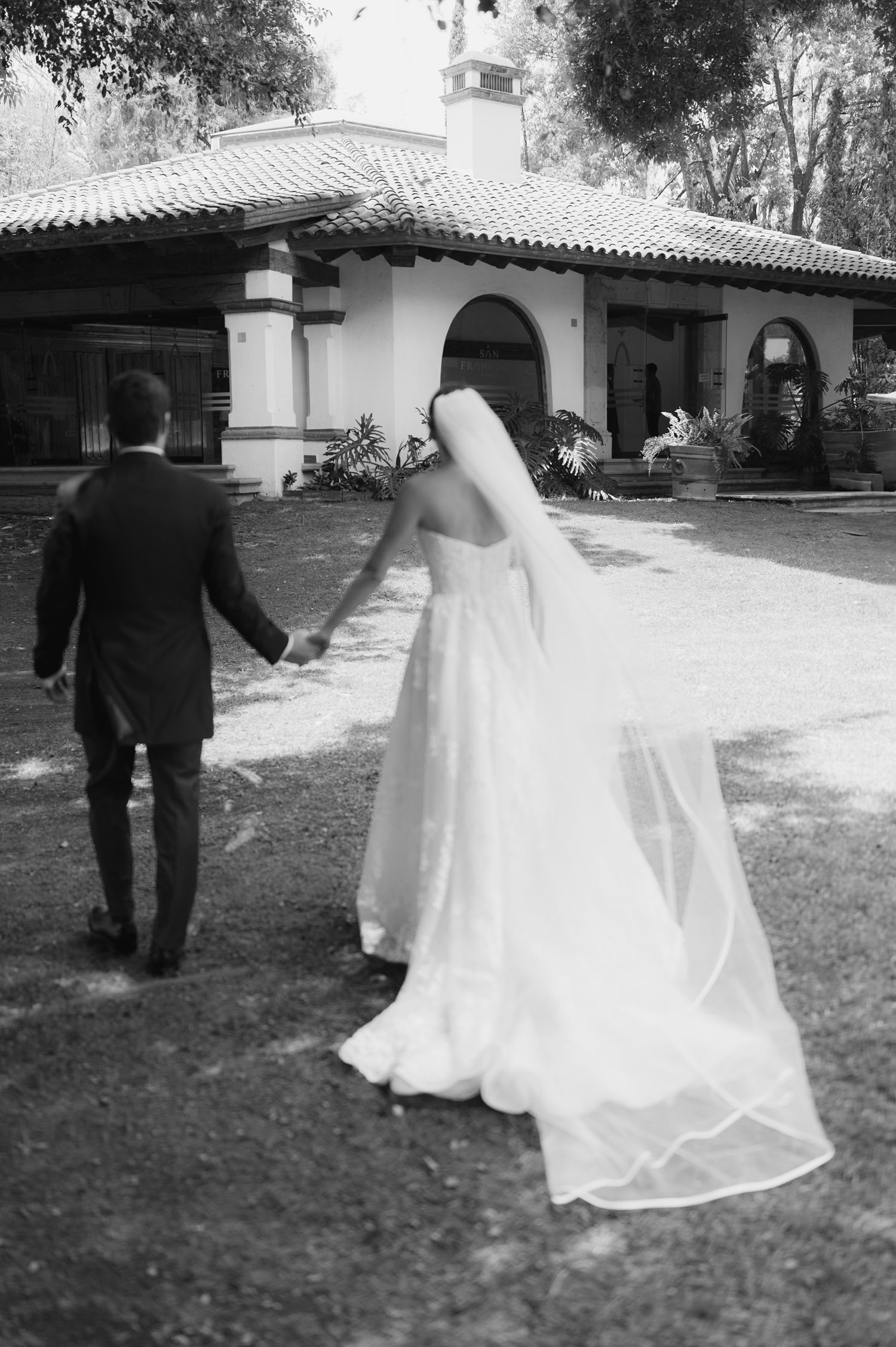 Black and white photo of bride and groom holding hands walking toward a Spanish-style venue.