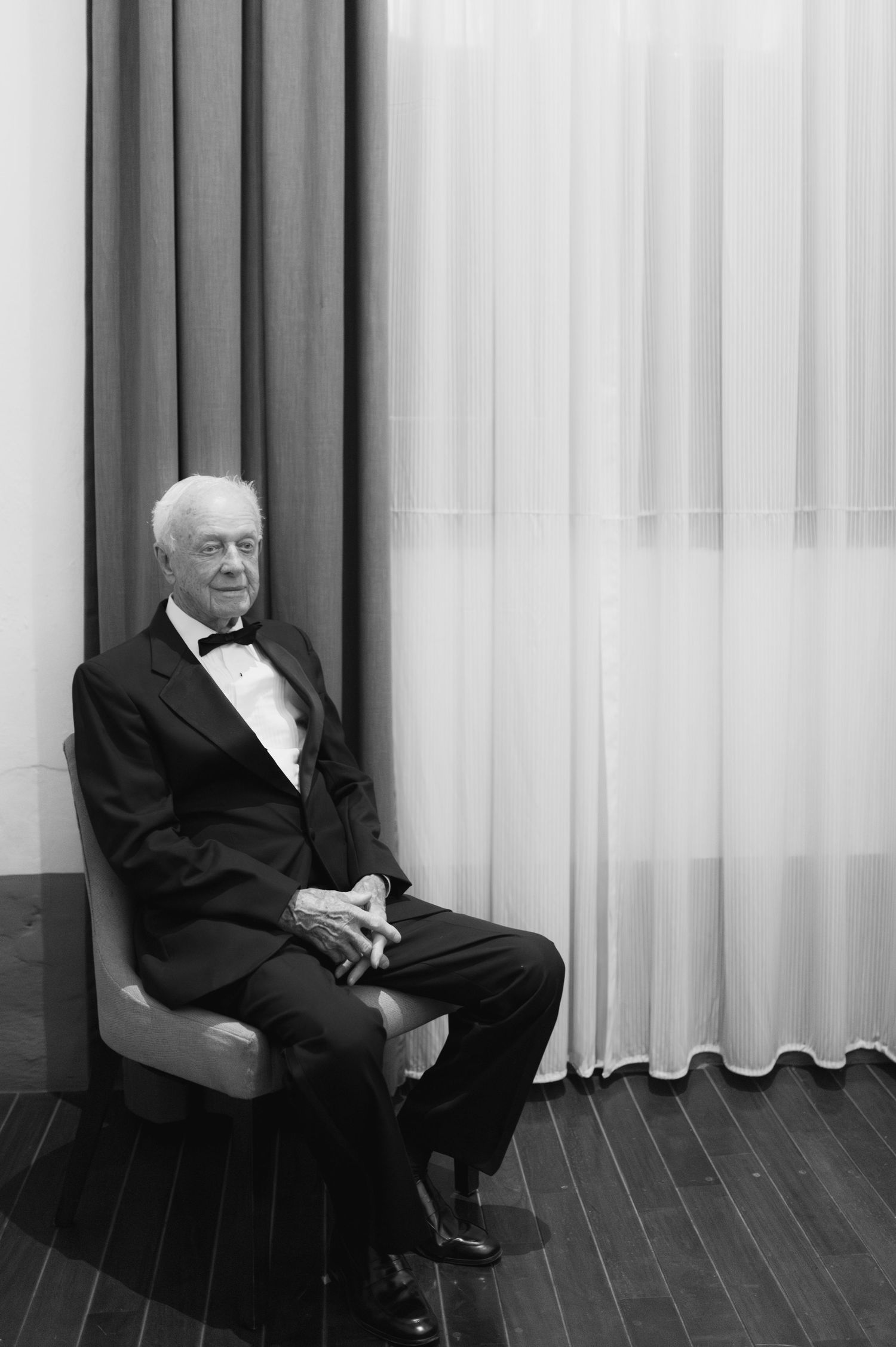 Elegant elderly man in a tuxedo seated in a chair by sheer curtains in black and white.