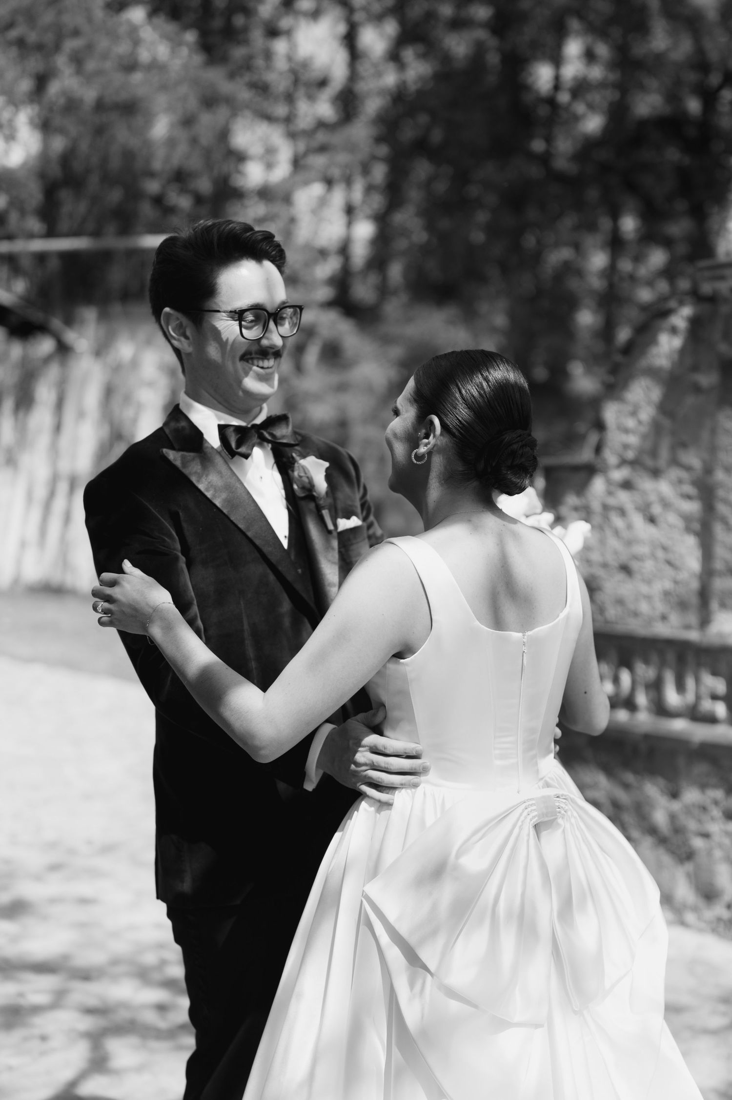 Black and white photo of smiling groom in bow tie and glasses embracing bride, showing back detail of wedding gown bow.