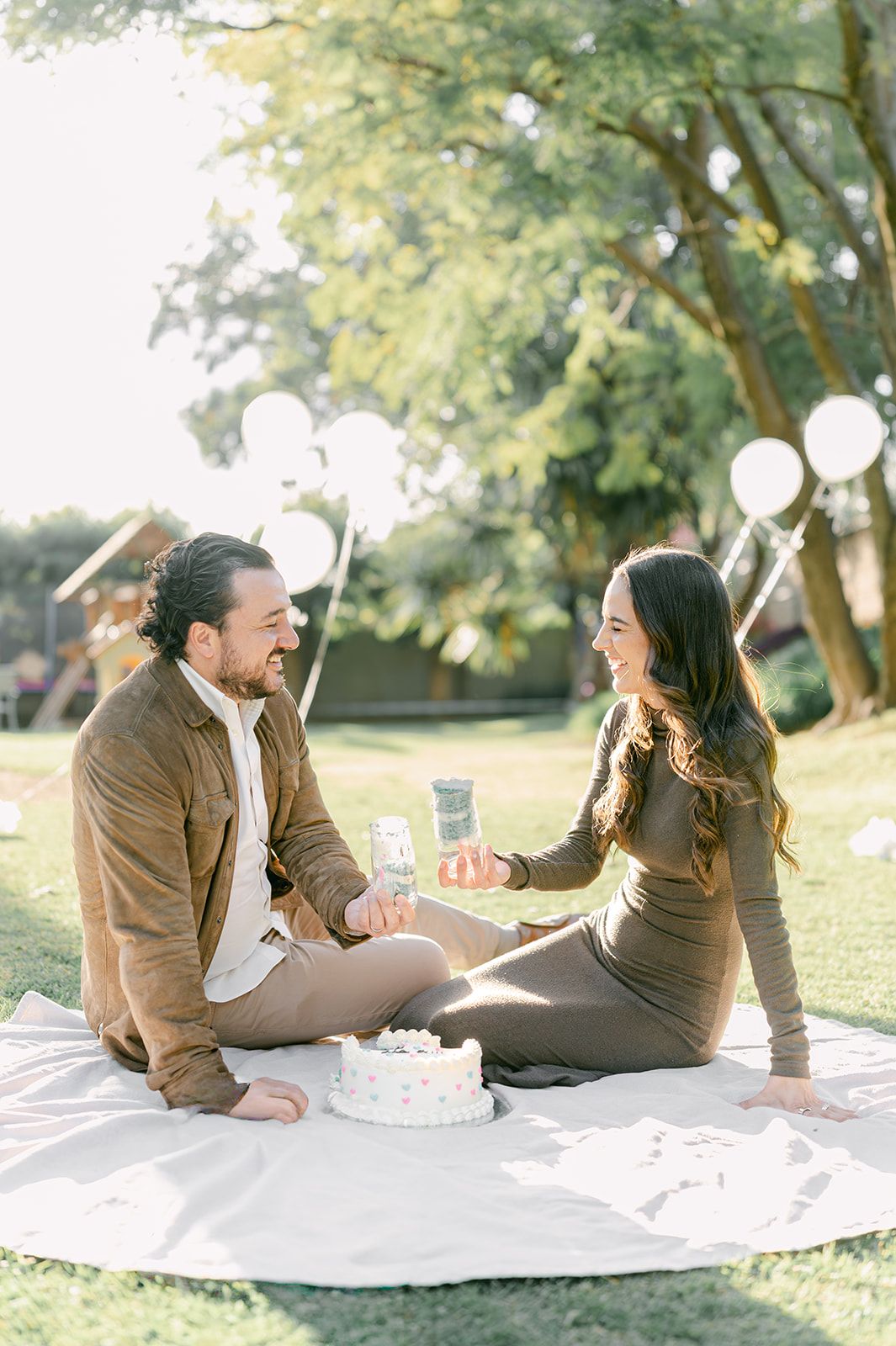 Happy couple sitting on white blanket in sunny park celebrating gender reveal with cake and drinks.
