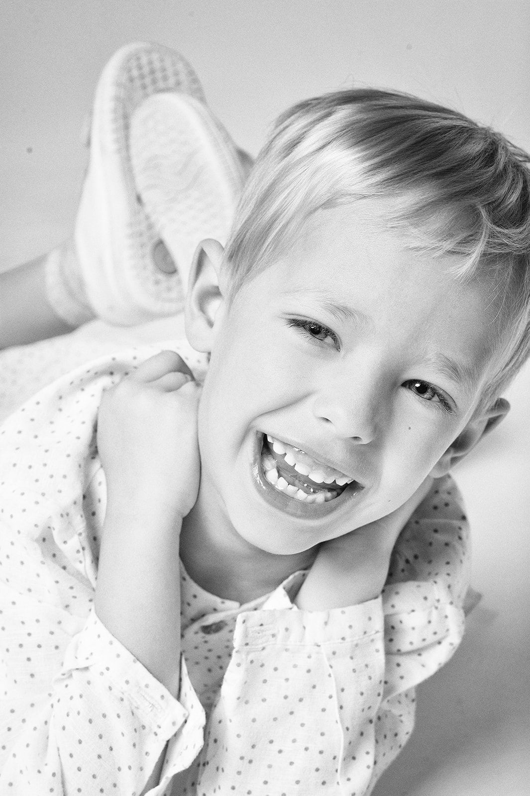 A laughing blonde child in a polka dot shirt poses playfully in a black and white studio portrait.
