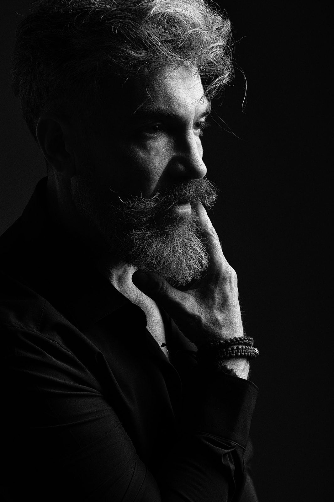 Black and white portrait of a bearded mature man in profile, resting chin on hand, wearing a bracelet.