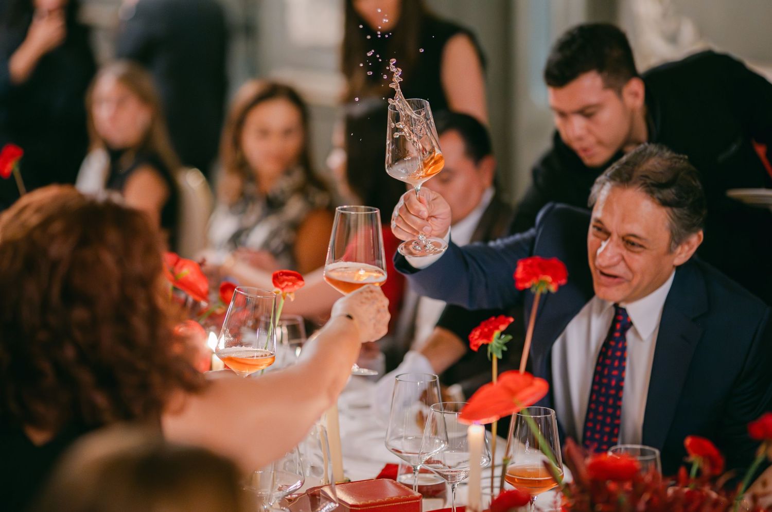 Guests raising wine glasses in a celebratory toast at an elegant dinner party with red floral centerpieces.