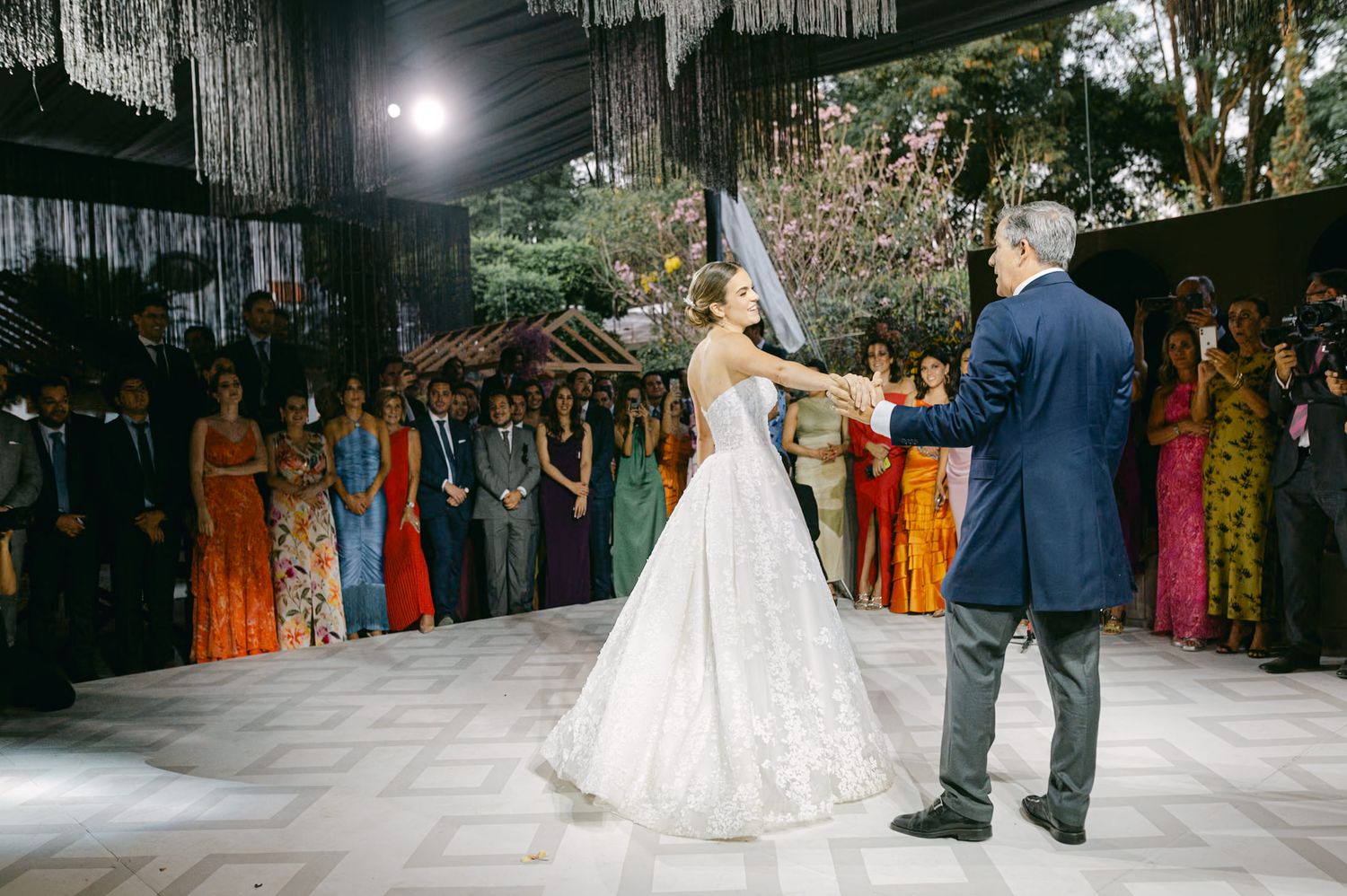 A wedding couple dances surrounded by colorfully dressed guests under elegant hanging decor.