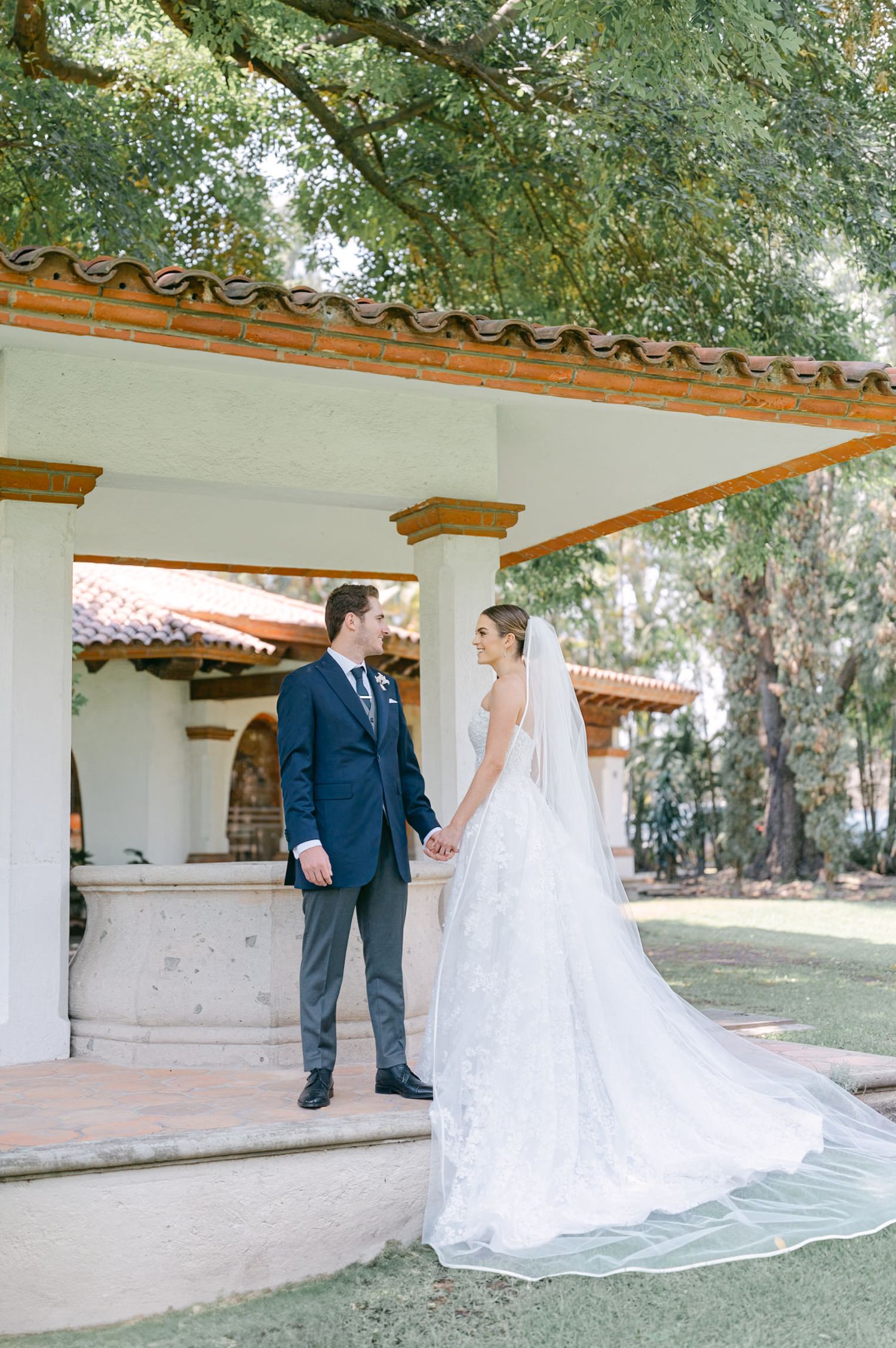 Bride and groom holding hands under ornate Spanish-style gazebo surrounded by lush green trees.