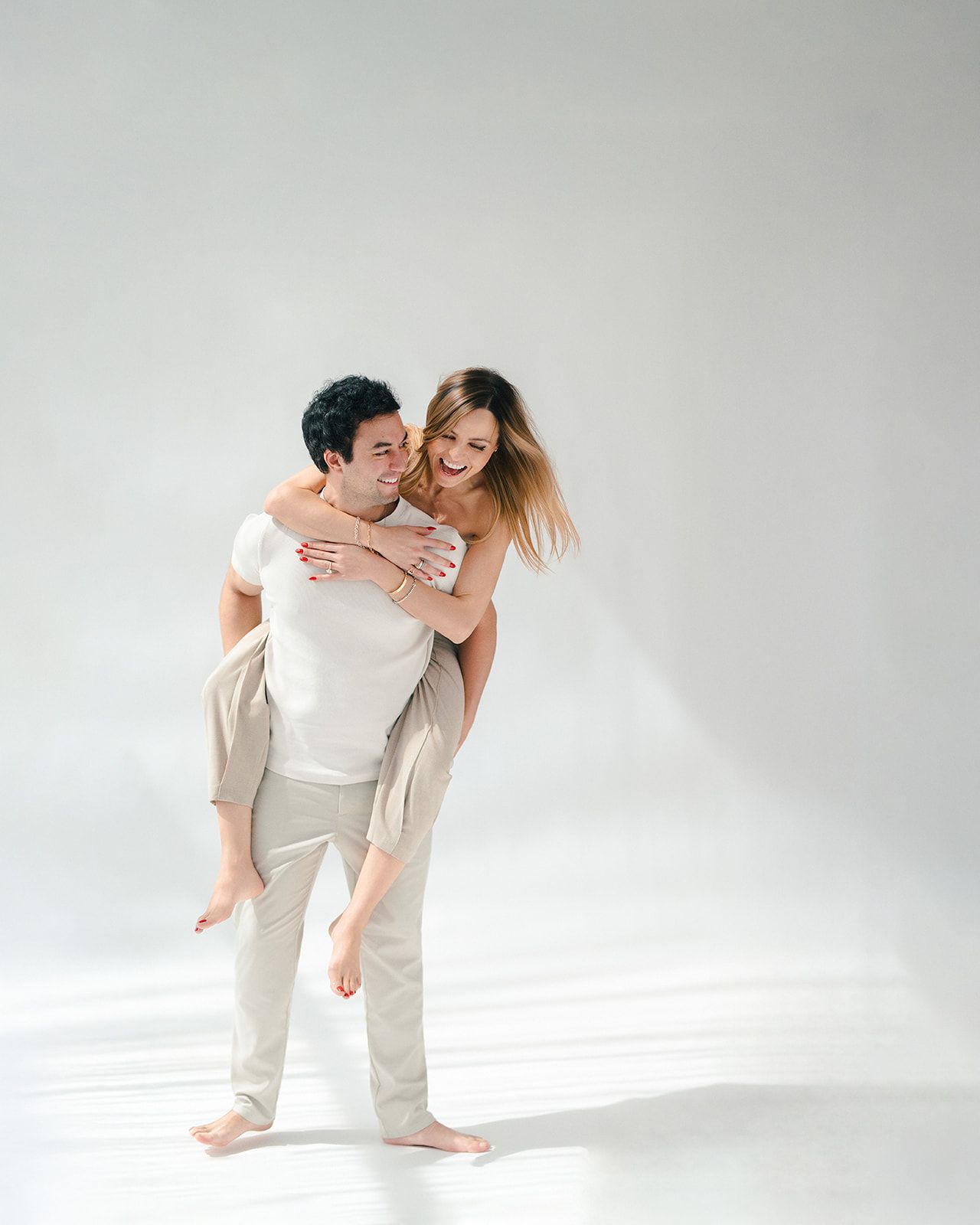 A man gives a woman a piggyback ride, both smiling and dressed in neutral tones against a white background.