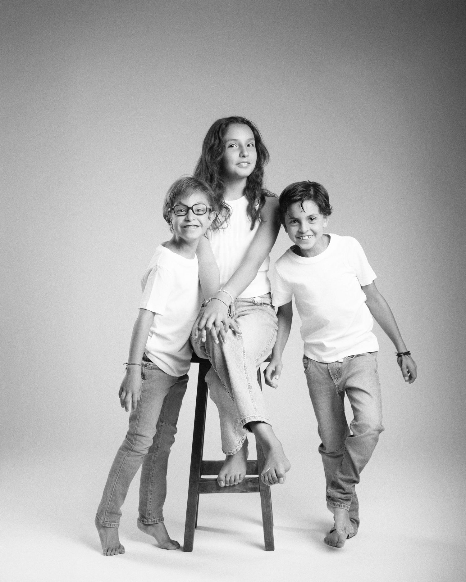 Black and white studio portrait of three children posing together with a stool.
