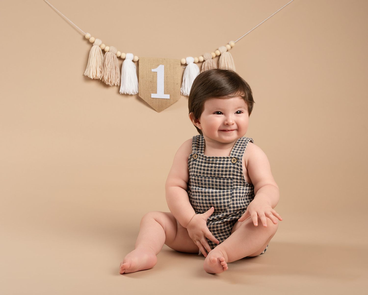 An adorable baby in a checkered romper smiles during a first birthday studio photo session.